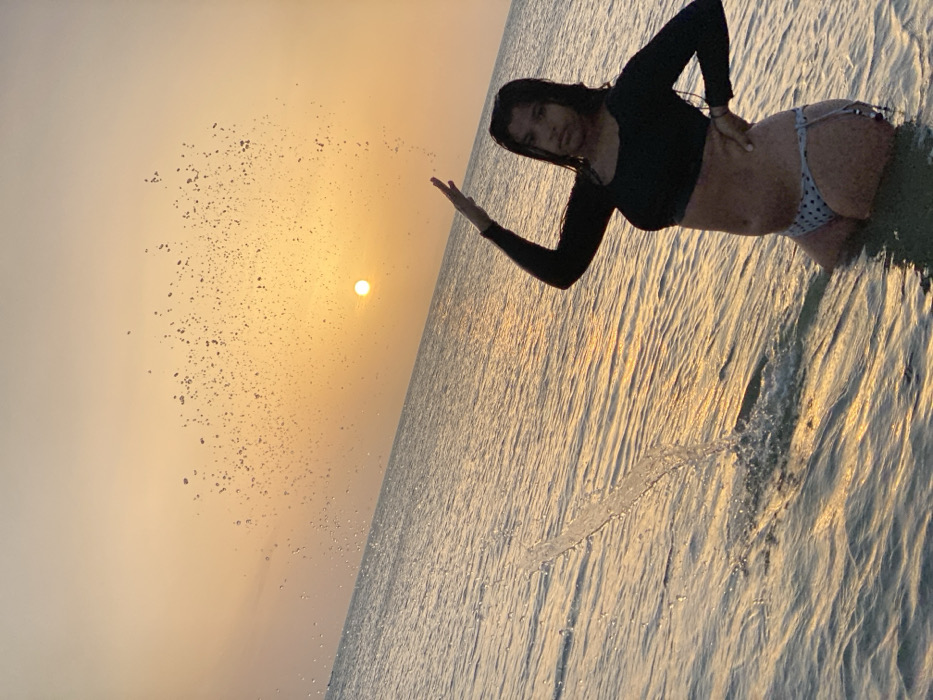 The image depicts a young adult female standing in the shallow waters of the sea at sunset in Juan Griego, Venezuela. The sky displays a clear golden-orange hue, with the bright, circular sun positioned low on the horizon. The calm water surface reflects the warm light of the sunset, creating golden streaks. The female, with dark, wet hair, wears a black long-sleeved crop top and light-colored, polka-dotted bikini bottoms. Her left arm is raised with the hand open and fingers pointing upwards, while her right hand rests on her hip. A visible stream of water originates from the lower-left foreground, arching upwards and to the right, dispersing into numerous illuminated droplets against the sunset sky.