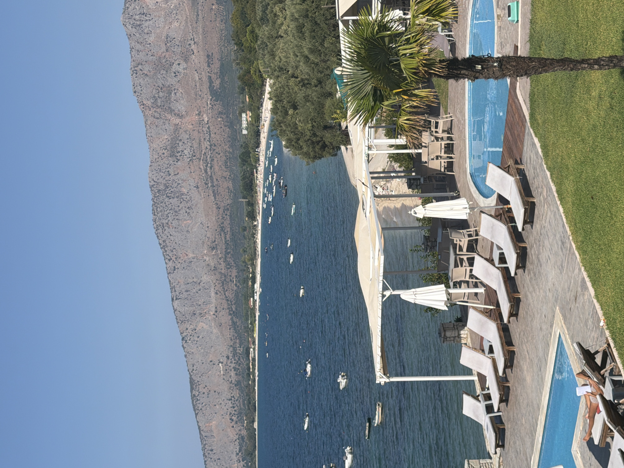 The image depicts a scenic view of the Municipal Unit of Alyzia, Greece. A tranquil blue sea stretches out towards a distant, rugged mountain range under a clear sky. Numerous small boats are scattered across the water. In the foreground, a resort area is visible, featuring a swimming pool, lounge chairs, umbrellas, and an outdoor dining area. A palm tree stands tall to the right of the pool. A person is lying on a lounge chair by the pool, reading a book. The resort is situated along a sandy beach lined with lush green trees.
