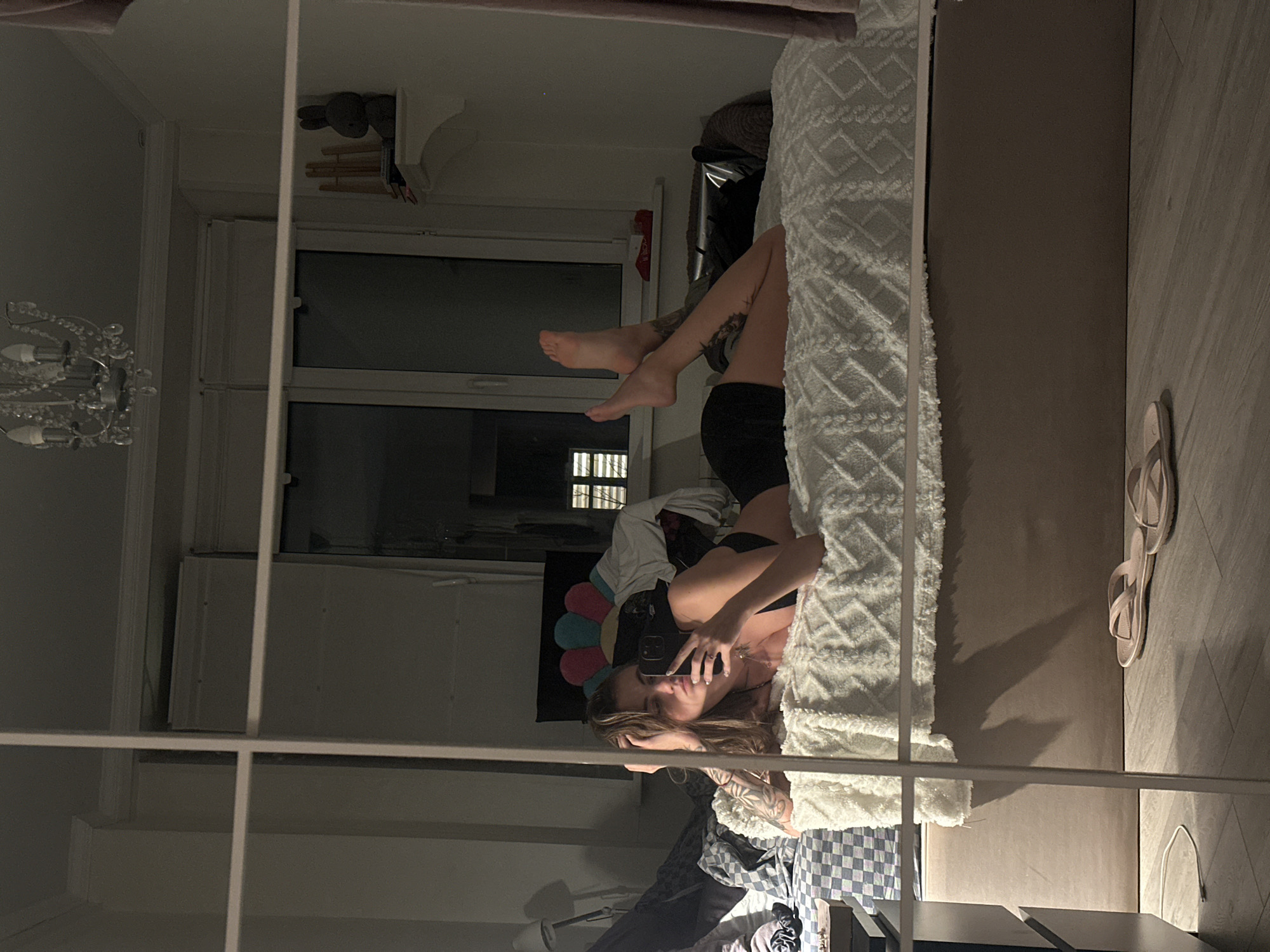 A young woman with light brown hair and tattoos on her arm is lying on a bed in a room, holding a smartphone to take a selfie in a mirrored closet door. She is wearing a black bra and black shorts. The bed is covered with a white textured comforter. In the background, a window, a chandelier, and a stuffed animal on a shelf are visible. The floor is made of light-colored wood, and a pair of pink sandals are on the floor. The overall lighting suggests it is evening.