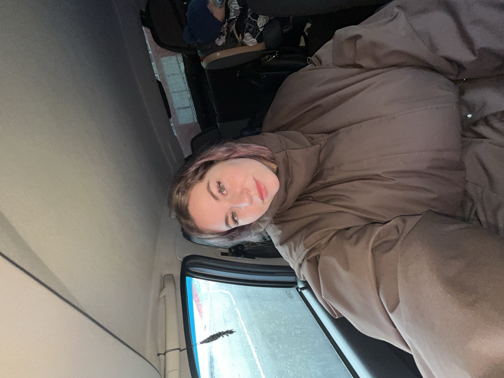 A young woman is in the back of a van, apparently sitting or reclining, wrapped in a large, brown puffer jacket. A window beside her is wet, indicating rain. Outside the window appears to be a street scene on an overcast day. Other items, including what appears to be luggage and a backpack, are also in the van. The location is Chaikovsky, Russia.