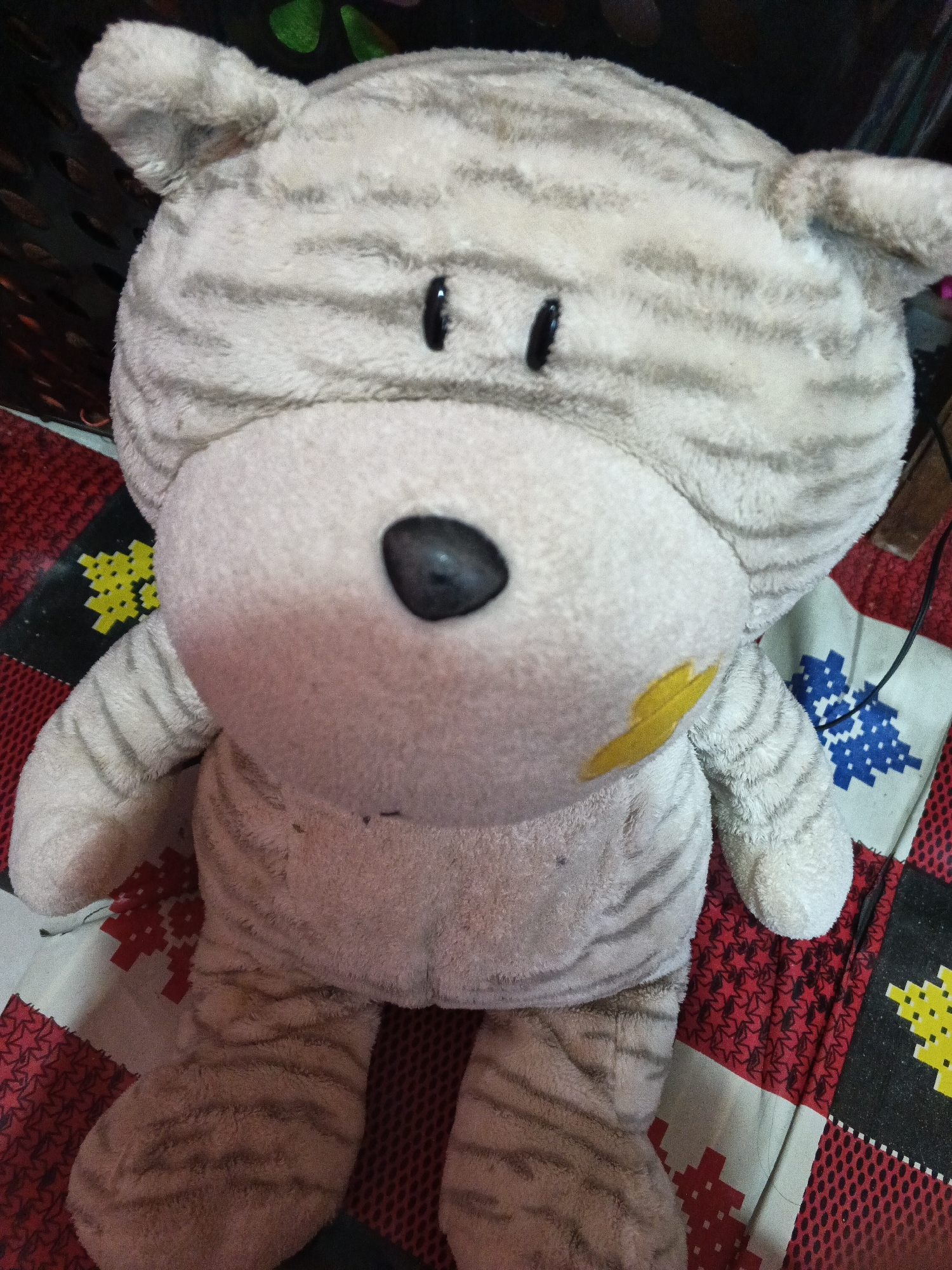 A close-up, eye-level shot focuses on a plush toy animal, resembling a cat, with grey and white stripes. The toy has large, round, black eyes, a black nose, and a light-colored snout with a yellow patch on the left cheek. It is positioned on a colorful, patterned surface with red, black, and white sections, featuring geometric designs and star shapes. The background is dark and out of focus, with some hints of green and brown. The lighting is somewhat dim, creating soft shadows. The image is taken indoors.