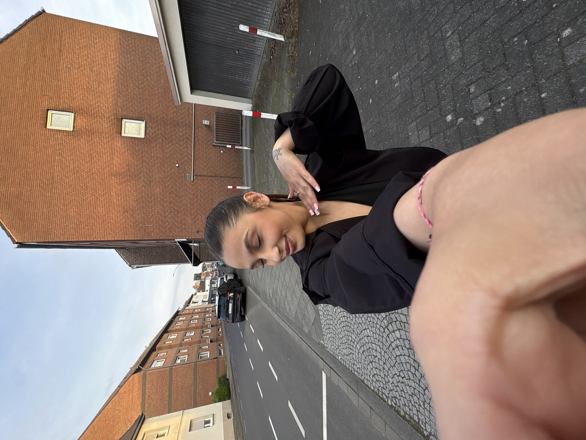 A woman is posing for a selfie on a street in Mönchengladbach, Germany. She is wearing a black top and has a bracelet on her wrist. A brick building and a garage are visible in the background. A street with parked cars and residential buildings extends into the distance. The sky is overcast.