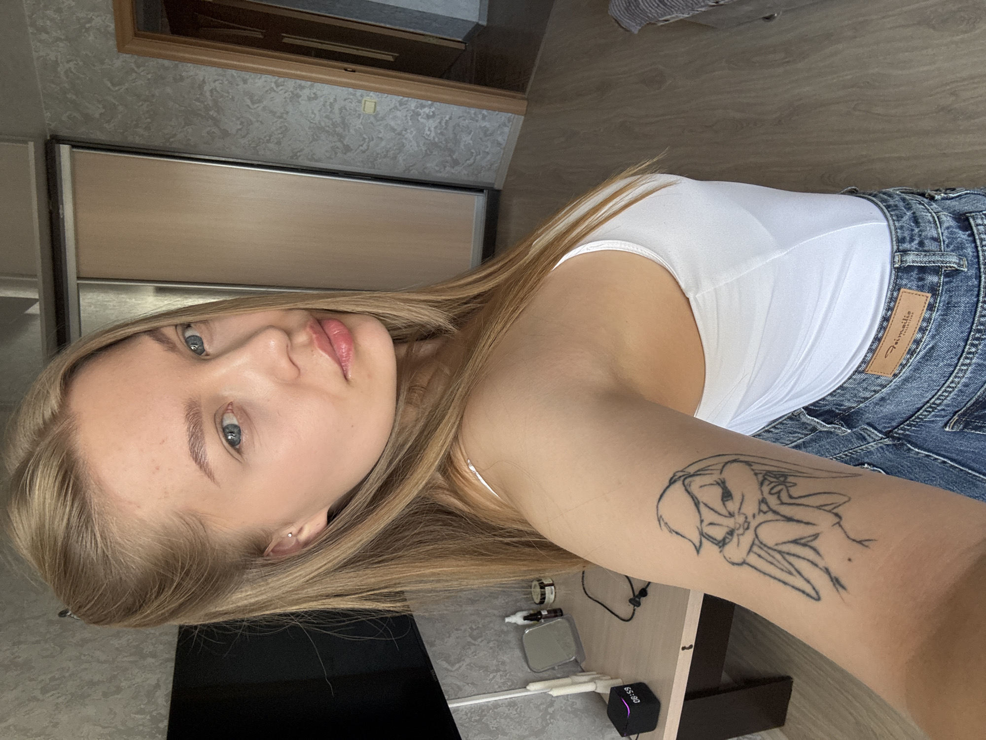 A fair-skinned young woman with blonde hair and blue eyes poses for a photo in what appears to be an apartment. She is wearing a white tank top and blue jeans. A tattoo of Bugs Bunny is visible on her left arm. A black digital clock displaying "08:59" is on a small desk next to her. Other items on the desk include a mirror, small bottles, and some tape. Behind her is a wall with light gray patterned wallpaper and a closet with sliding doors. The floor is made of light brown wood. It is not possible to definitively confirm the location of Chaikovsky, Russia from the image, however.
