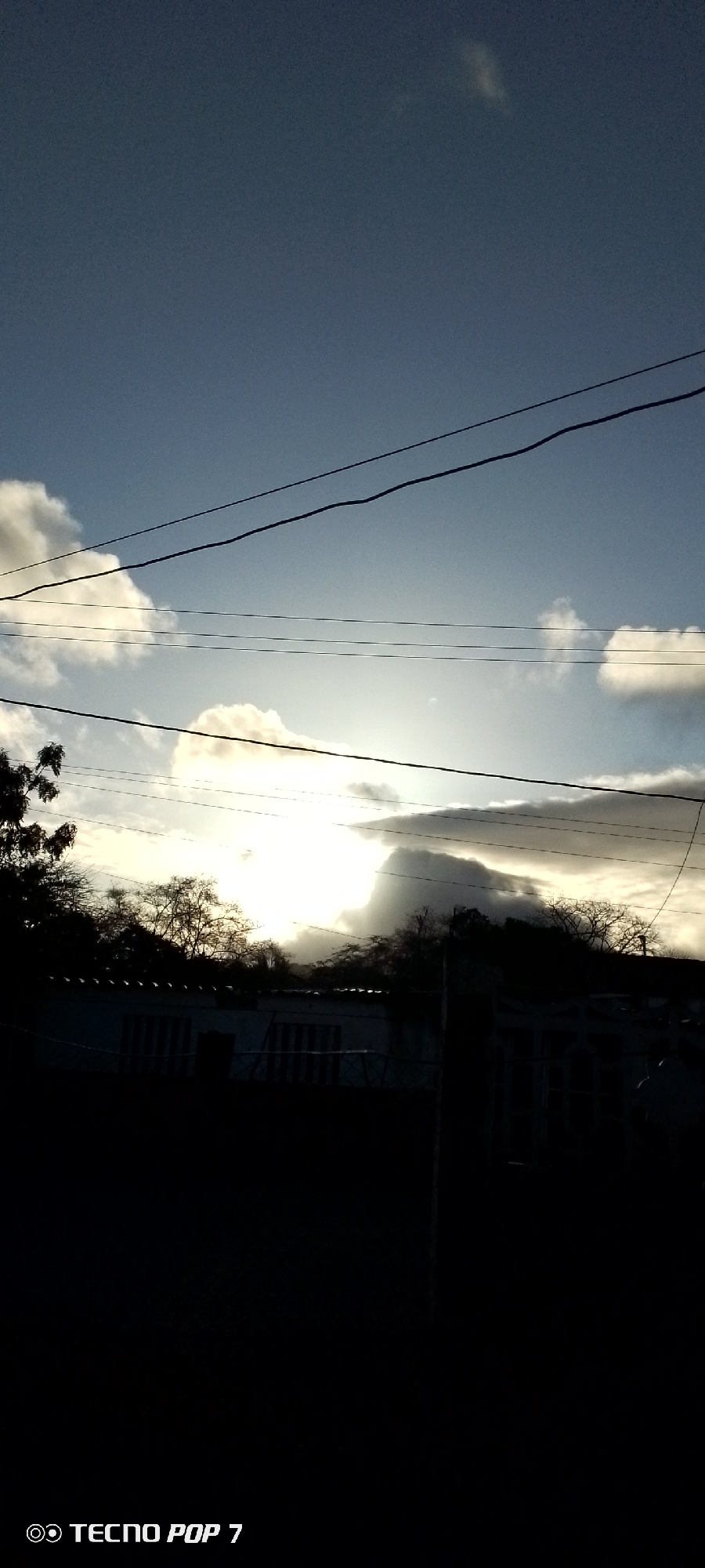 The image depicts a landscape view in Altagracia, Venezuela. The sky is a mix of blue and white, with clouds scattered throughout. Power lines stretch across the upper portion of the frame. The sun is obscured by clouds, creating a bright glow. In the midground, there are trees and what appears to be a building. The foreground is mostly dark, obscuring details. The TECNO POP 7 watermark is visible in the bottom left corner.