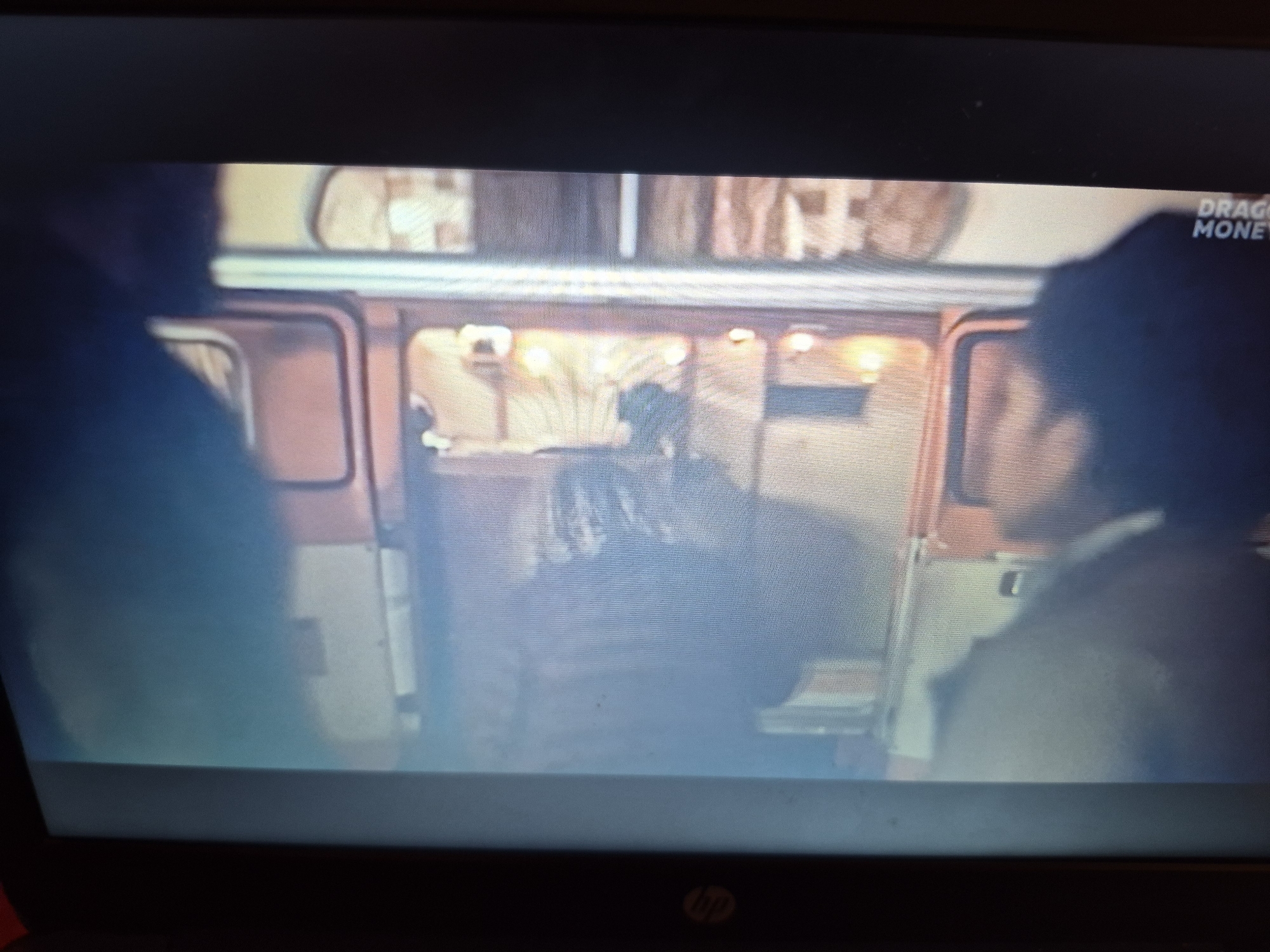 A television screen displays a scene from what appears to be a documentary or reality show, indicated by the text "DRAGO MONEY" in the upper right corner. The setting seems to be an interior, possibly a train carriage or a mobile unit, with warm lighting emanating from within. In the foreground, the silhouettes of two individuals are visible, one on the left and one on the right, looking into the illuminated interior. Inside, a person is seated, facing away from the camera, with their hands on their lap. The overall image is slightly blurry, suggesting it was captured from a distance or in low light conditions. No specific location in Sandwell, United Kingdom, can be identified from the visual information provided.