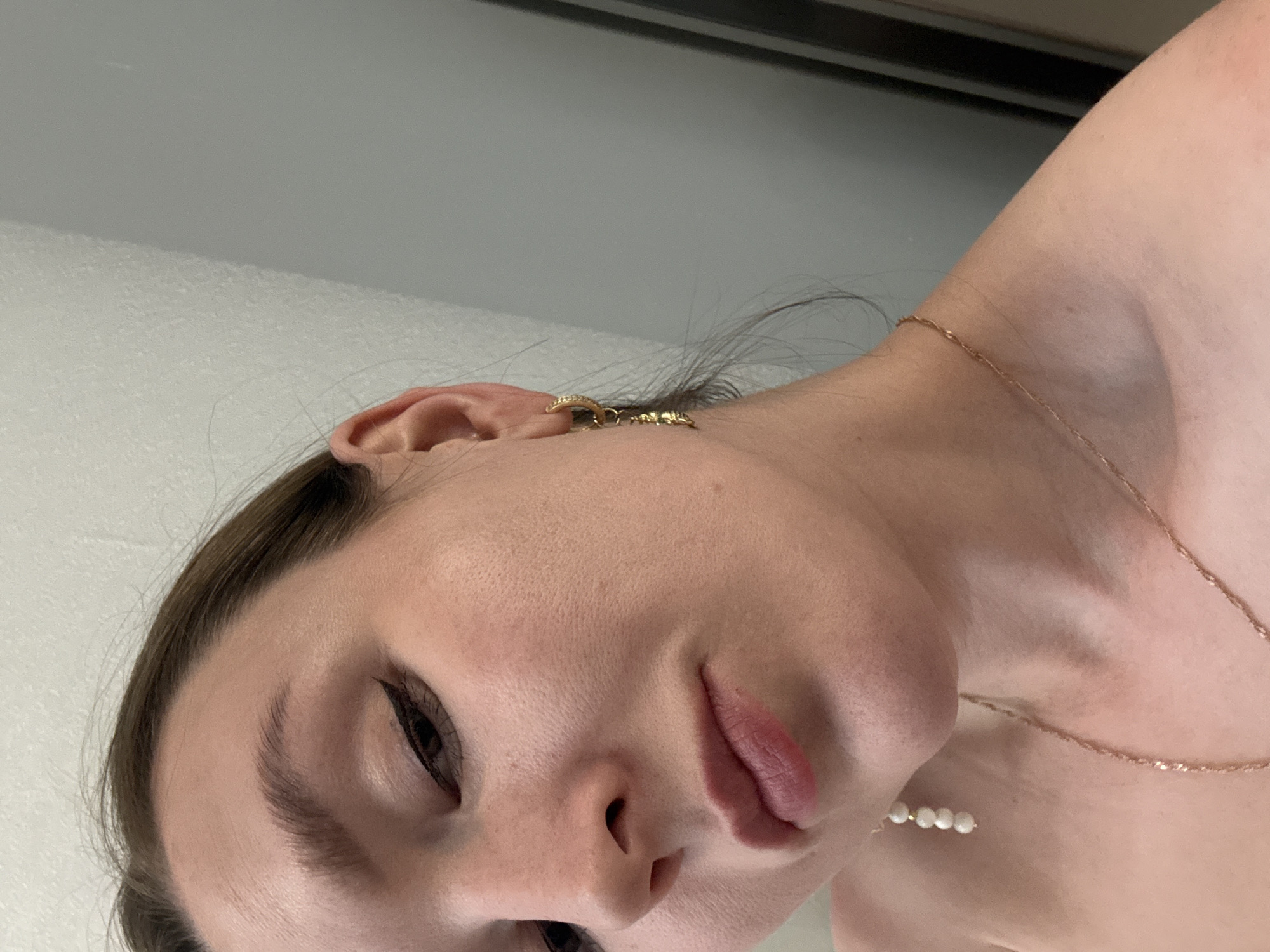 A close-up, rotated portrait of a person, likely a woman, taken in Izhevsk, Russia. The image is oriented sideways, with the subject's head tilted sharply to the right. She has fair skin, dark brown hair partially visible, and is wearing makeup, including winged eyeliner and pinkish lip color on her slightly parted lips. Her expression is neutral and relaxed.
Jewelry is prominently featured. She wears a gold-colored hoop earring with textured details in her visible ear. A thin, gold-colored chain necklace with a textured design is draped around her neck and collarbone. Near her chest, a cluster of small, white, pearl-like beads is visible, possibly part of the necklace or a separate accessory. Another gold-toned dangle earring, also featuring small white, pearl-like beads, hangs near her left shoulder.
The background is simple, consisting of light-colored, possibly white and grey, walls or surfaces, indicating an indoor setting. The lighting is soft and even. No text is visible in the media file.