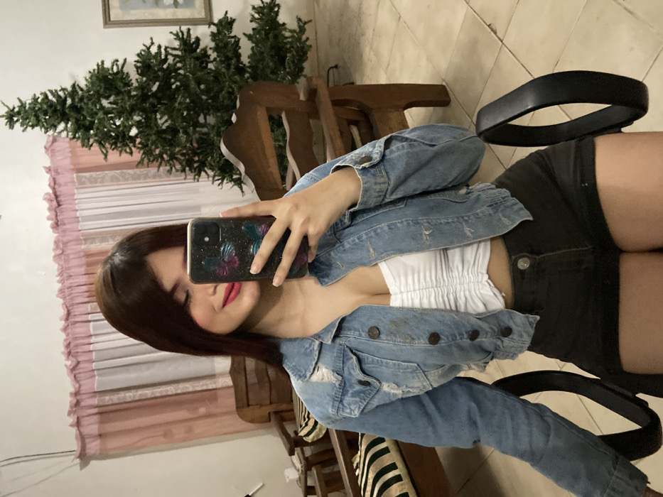 A woman captures a selfie in a mirror, revealing a tilted, mid-angle shot. She wears a cropped white top, a light blue denim jacket, and dark shorts, seated on a chair with a black armrest. Her brown hair frames her face, and she holds a smartphone with a patterned case. In the background, a faux Christmas tree and partially draped curtains add to the domestic setting. The tiled floor and wooden furniture suggest an indoor environment.