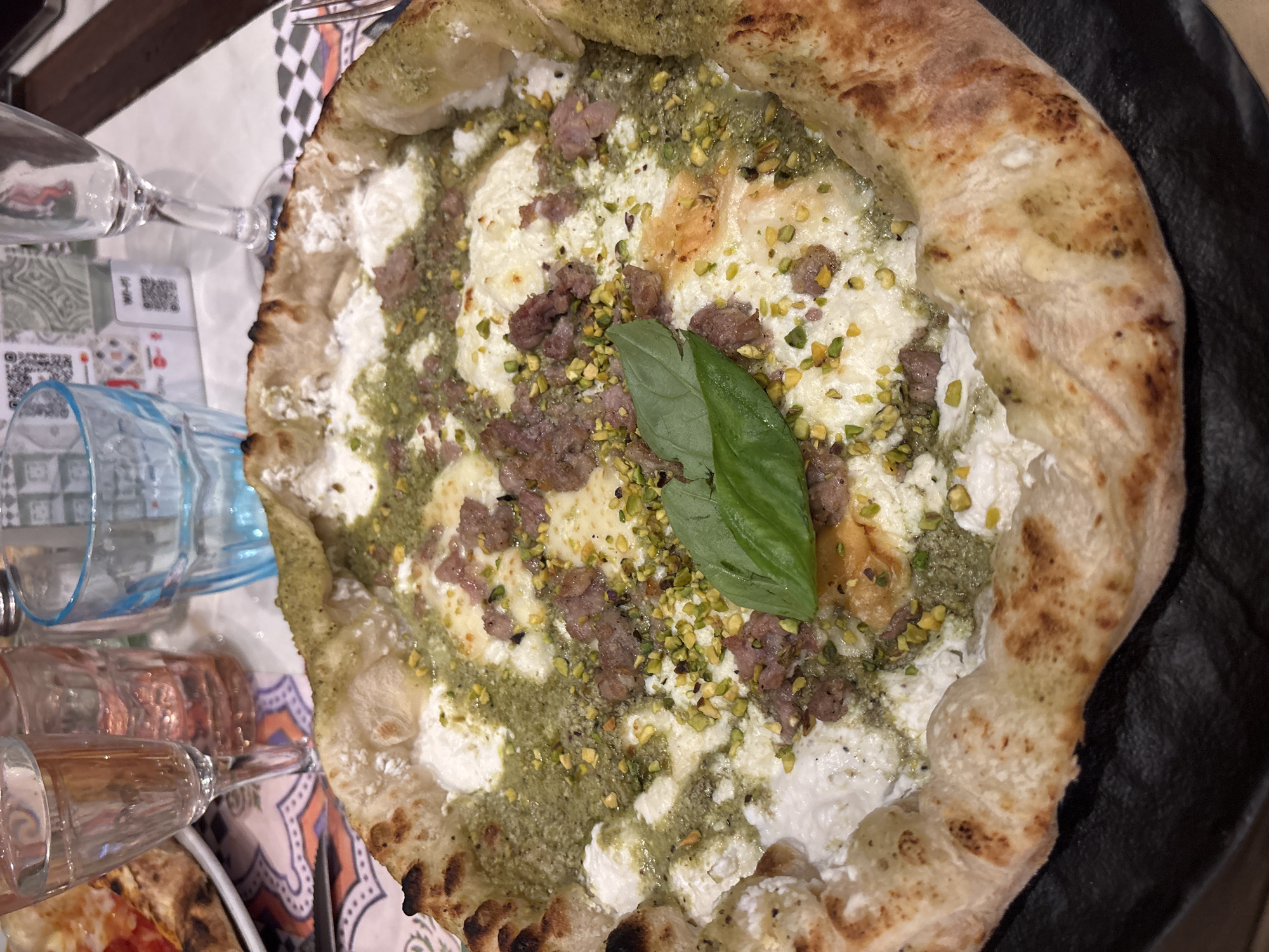 A close-up, top-down shot captures a gourmet pizza with a puffy crust, glistening with a green sauce, topped with white cheese, crumbled sausage, and chopped pistachios. A fresh basil leaf rests in the center. In the background, out of focus, are two glasses of water and another pizza on a decorative napkin. This scene is consistent with a meal at a restaurant in Milan, Italy, known for its culinary scene and stylish ambiance.