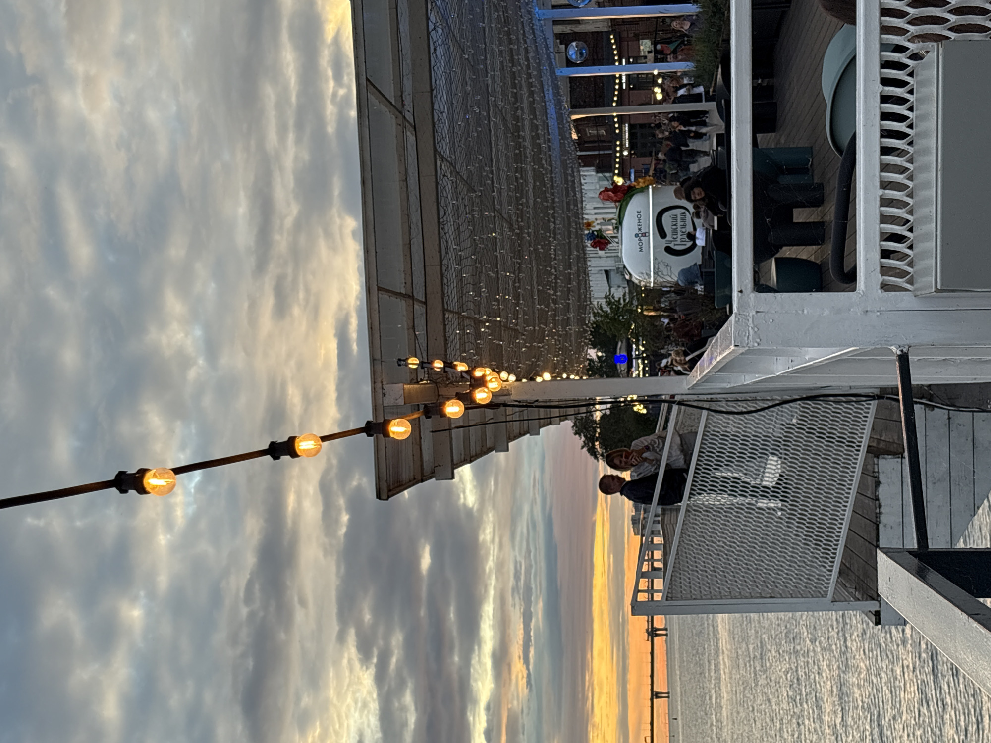 The image captures a waterfront scene at dusk in Saint Petersburg, Russia. A couple stands on a white railing overlooking the water, engaged in conversation. A string of illuminated bulbs stretches across the top of the frame, casting a warm glow against the cloudy sky, which is tinged with orange and yellow from the setting sun. In the background, a white food truck with Russian text, likely selling ice cream, is visible, along with other patrons enjoying the evening. The overall atmosphere suggests a lively outdoor gathering place by the water.