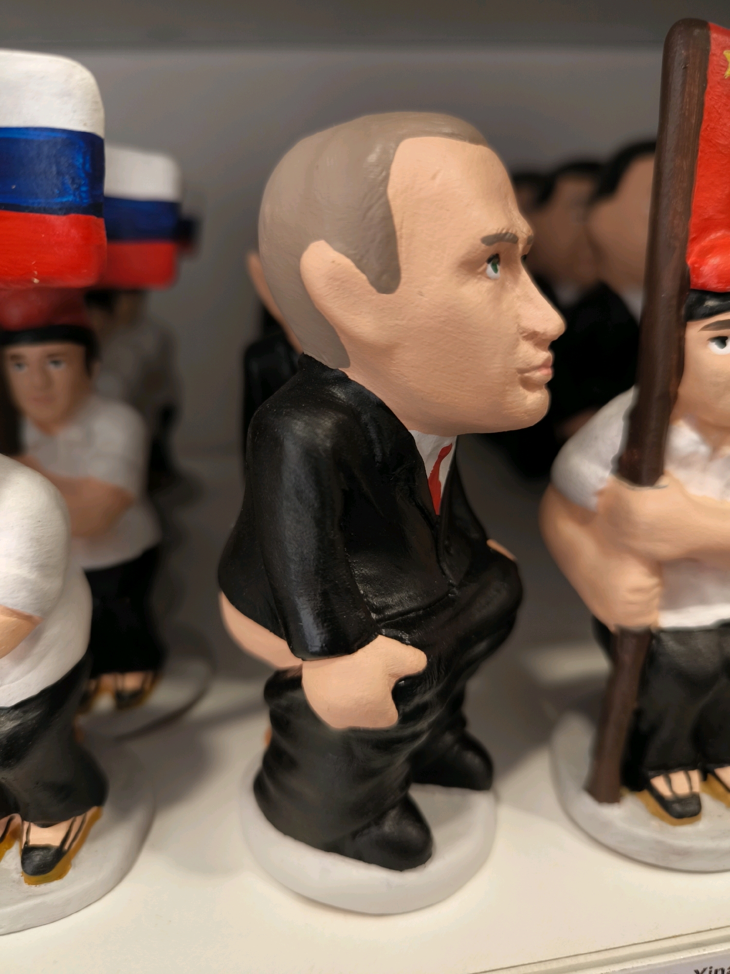 The image shows a close-up of a row of novelty figurines. The most prominent figurine in the foreground is a caricature of Vladimir Putin, depicted in a black suit with his pants pulled down to his ankles. He is facing to the right in profile, with his head turned slightly upwards and to the right, revealing his facial features and a hint of green in his eye. His left hand is visible, holding the fabric of his trousers. The figurine is standing on a white base.
Behind the Putin figurine, other similar figurines are visible, though they are out of focus. To the left, a figurine with a red, white, and blue flag (resembling the Russian flag) is partially visible. To the right, another figurine is holding a dark brown pole with a red flag attached. The background is a plain, light gray surface.
The figurines appear to be made of a painted material, possibly ceramic or resin, with a matte finish. The style is cartoonish and exaggerated. The overall impression is of a display of political satire or novelty items. The question asks for the location in the city of Bilbao, Spain, but there are no specific visual cues in the image to indicate this location.