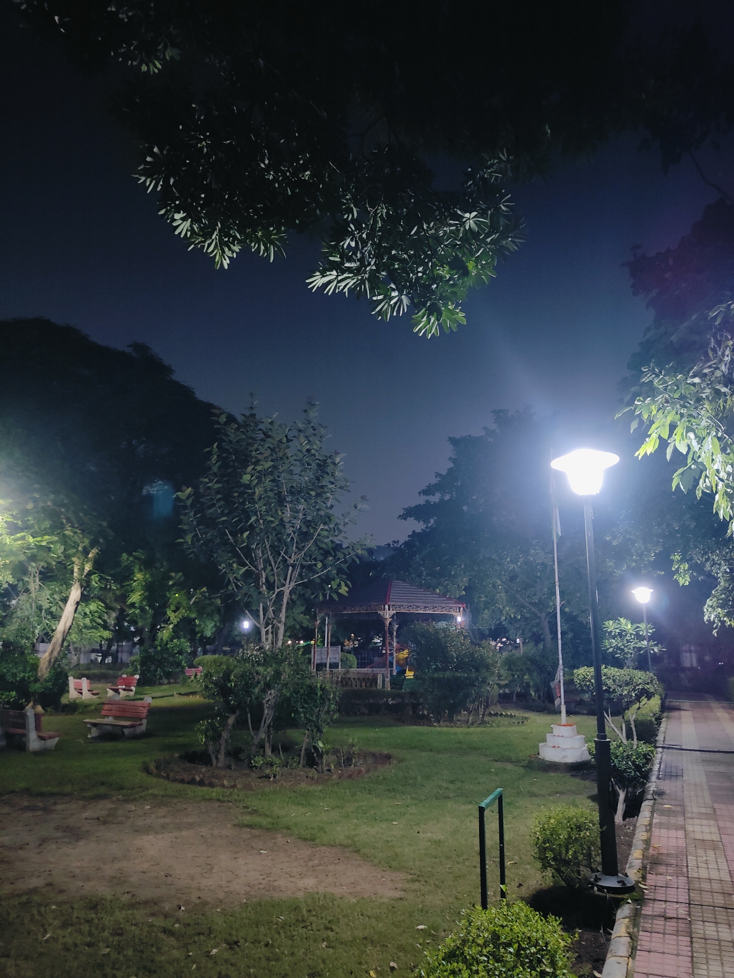 This image depicts a park at night in Gurgaon, India. The scene is illuminated by streetlights, casting a glow on the manicured lawns, pathways, and surrounding trees. Several red and white benches are scattered throughout the park, some empty, others occupied by a few individuals. A gazebo-like structure is visible in the background, suggesting a common area for gatherings or relaxation. The overall atmosphere is tranquil, with the darkness of the night contrasting with the bright lights, creating a sense of calm and serenity. The presence of lush greenery indicates that the park is well-maintained and offers a natural escape within the urban environment.