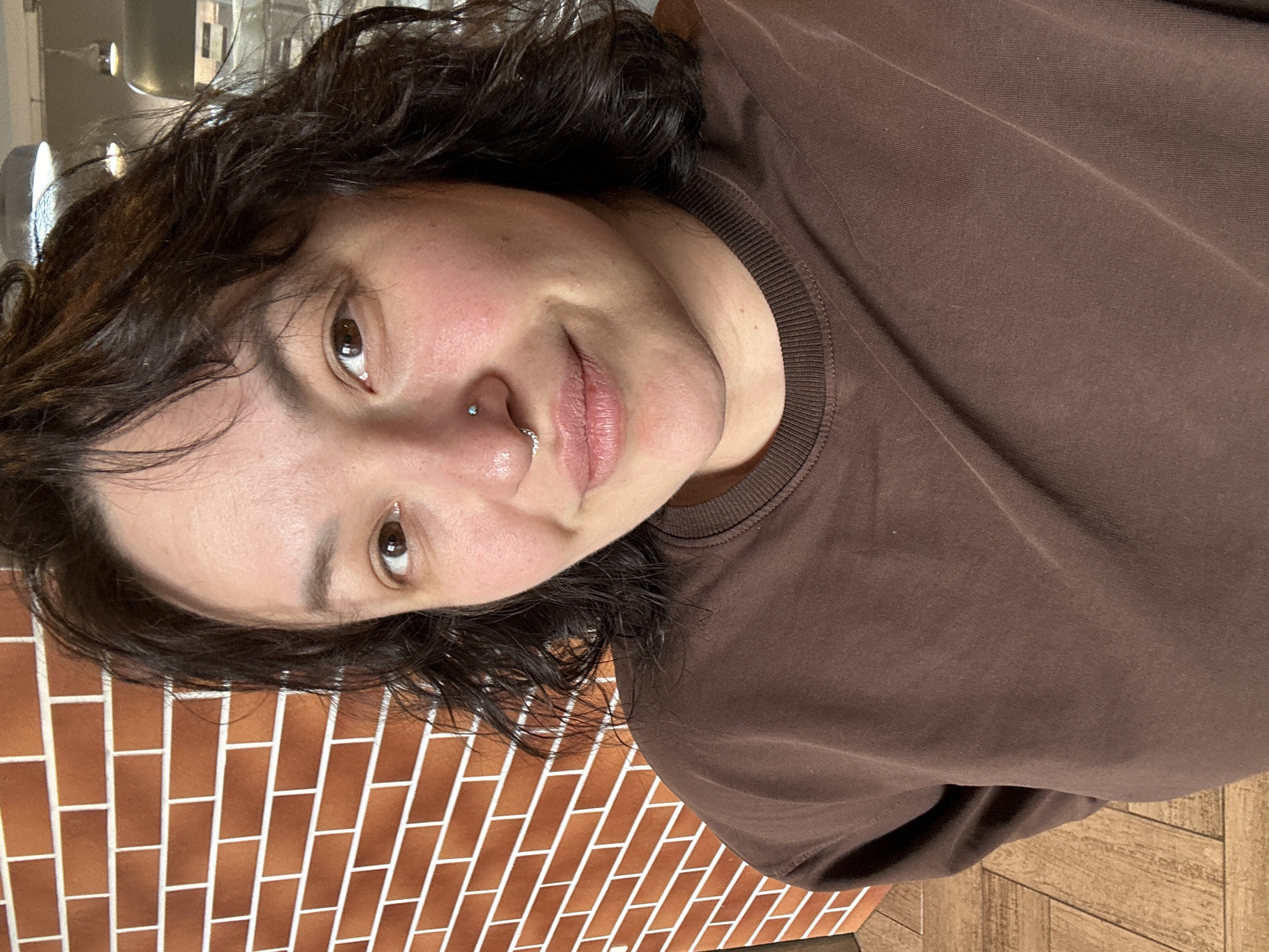 A selfie of a person with dark, wavy hair, light skin, and brown eyes. They have a nose piercing on the left side and are wearing a brown t-shirt. The background consists of an orange brick wall on the left and a light brown wooden surface on the bottom right. The photo's location is indeterminable from the provided image and without additional information, it's impossible to definitively say it's in Votkinsk, Russia.