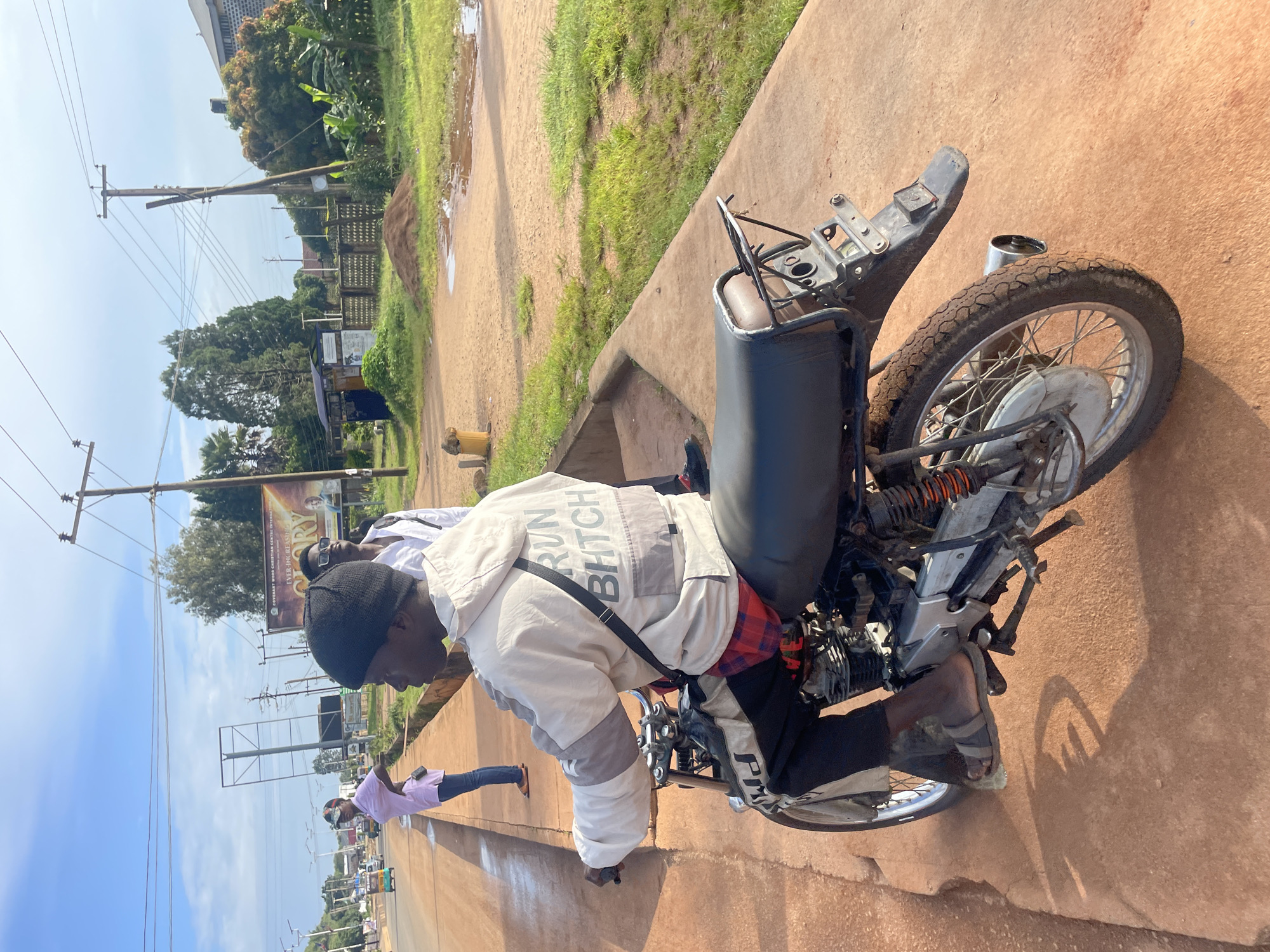 The image depicts an outdoor scene on a sunny day in Jos, Nigeria, showing a man sitting on a motorcycle on a reddish-brown paved surface. The man is wearing a black beanie, a light-colored jacket with gray accents, and black pants. The jacket has the words "RUN BHITCH" printed in large gray letters on the back. He is also wearing plaid shorts or a plaid undershirt visible at the waist. On his feet, he has grey and black sandals. The motorcycle is dark-colored with visible mechanical parts and orange suspension springs. The rear of the motorcycle shows a tail rack and a license plate holder.
In the background, to the left, a person in a pink shirt and blue jeans is walking away from the camera, and a white vehicle is visible on a road. Further in the background, there are utility poles with wires, trees, and buildings, including a large billboard with text and images. To the right of the motorcycle, there is a grassy area with some puddles of water and a pile of dirt. The overall environment suggests a street or roadside in a developed area of Jos.