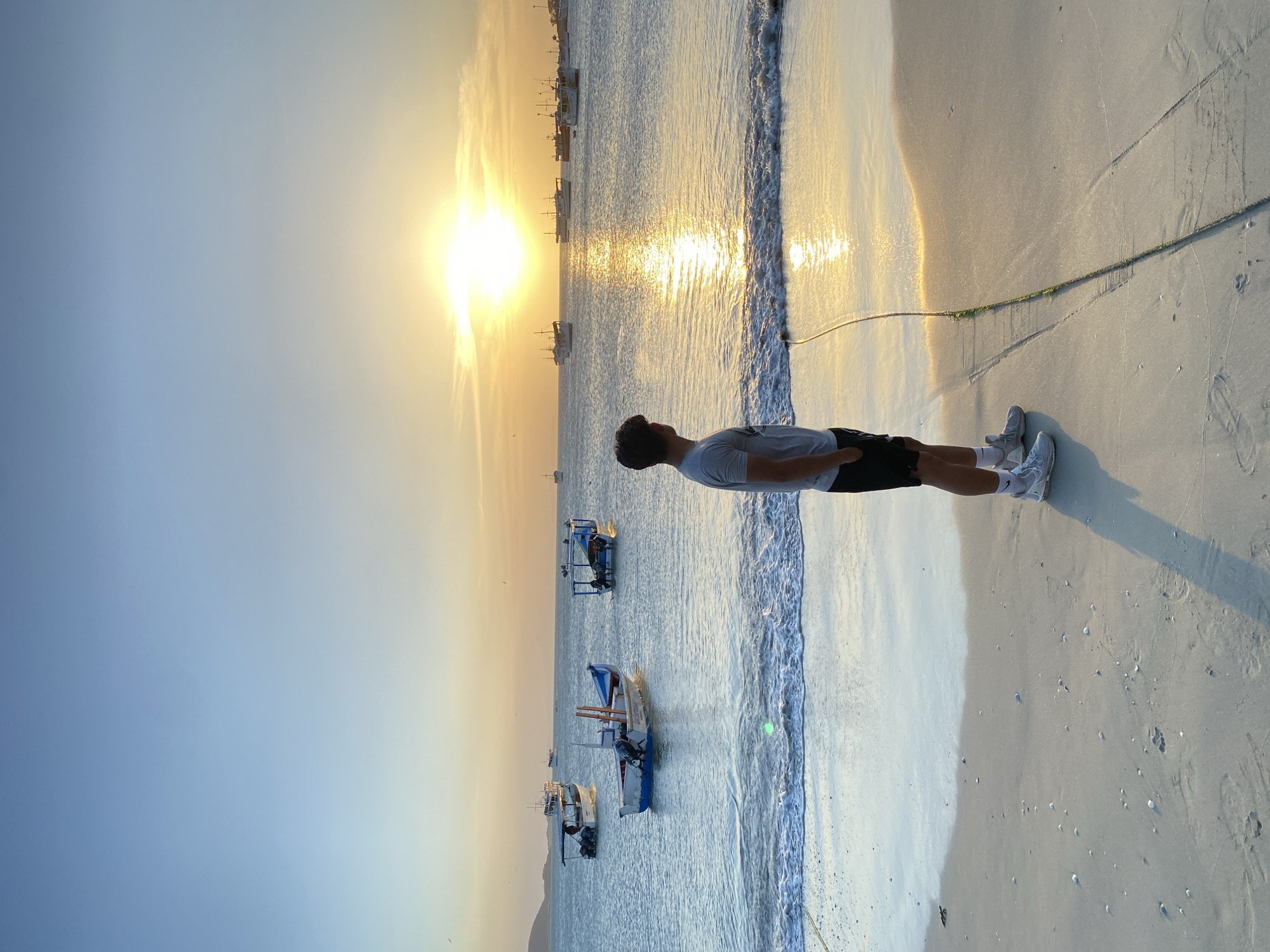 A vertical, eye-level shot captures a man standing on a sandy beach in Juan Griego, Venezuela. He is facing the sea with his back to the camera. The man is wearing a light-colored t-shirt, dark shorts, and white shoes with white socks. To his right, a rope stretches along the sand towards the water. The sea is calm with gentle waves breaking on the shore, reflecting the light from the setting sun. Several small fishing boats are visible in the water. The sky transitions from a soft blue to a golden yellow near the horizon, where the sun is setting, casting a warm glow over the scene.