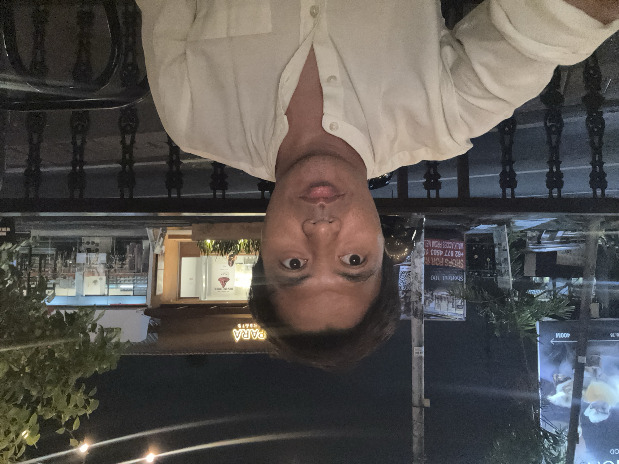 A man in a white, collared, button-up shirt is captured in a frontal, close-up shot. The background suggests an outdoor evening setting, possibly a restaurant or cafe, with a building displaying a neon sign that reads "PARA REATS". String lights are visible overhead, and greenery from a tree is present on the right side of the frame. Behind the man, a black metal fence or railing separates the viewer from a street or pathway, with streetlights and a dark road visible. Further in the background, a large billboard advertising "MOOD" is partially obscured by a pole. There's a sign with text including "Shortcut 300" and a phone number. The overall lighting indicates it is nighttime.