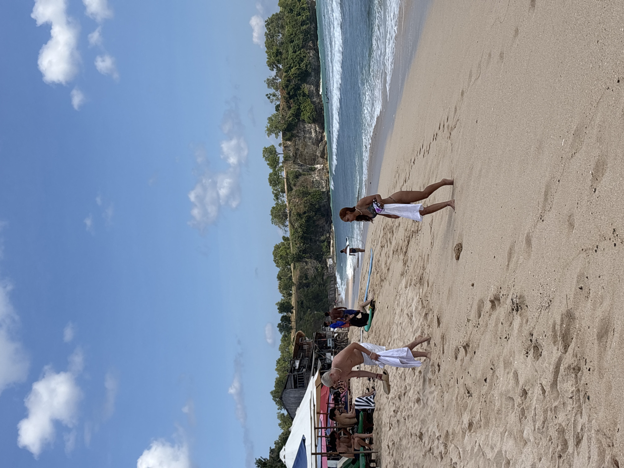 This image captures a wide-angle view of a beach in Kuta Selatan, Indonesia, under a clear blue sky with scattered white cumulus clouds. The foreground consists of light golden-brown sand marked with numerous footprints.
In the mid-ground, a man with light-colored hair and a necklace, wearing white swim shorts, is depicted bending slightly. He holds a white towel in his left hand and a pair of light-colored sandals in his right. To his right, a woman with reddish-brown hair, dressed in a dark bikini, walks barefoot toward the right edge of the frame, holding a white towel.
To the far left of the frame, under large white and blue beach umbrellas, several individuals are seated on lounge chairs. Further back, near the base of a cliff, various wooden structures resembling beach shacks or cafes are visible amidst trees.
The ocean, a blue-green hue, occupies the right side of the frame, exhibiting breaking waves. Multiple small figures, presumed to be surfers, are visible in the water and near the shore with surfboards. The background is dominated by a steep, vegetated cliff face topped with dense green foliage, extending along the coastline. One person in the mid-ground right is seen carrying a surfboard towards the water, while another is seated on a surfboard on the sand.