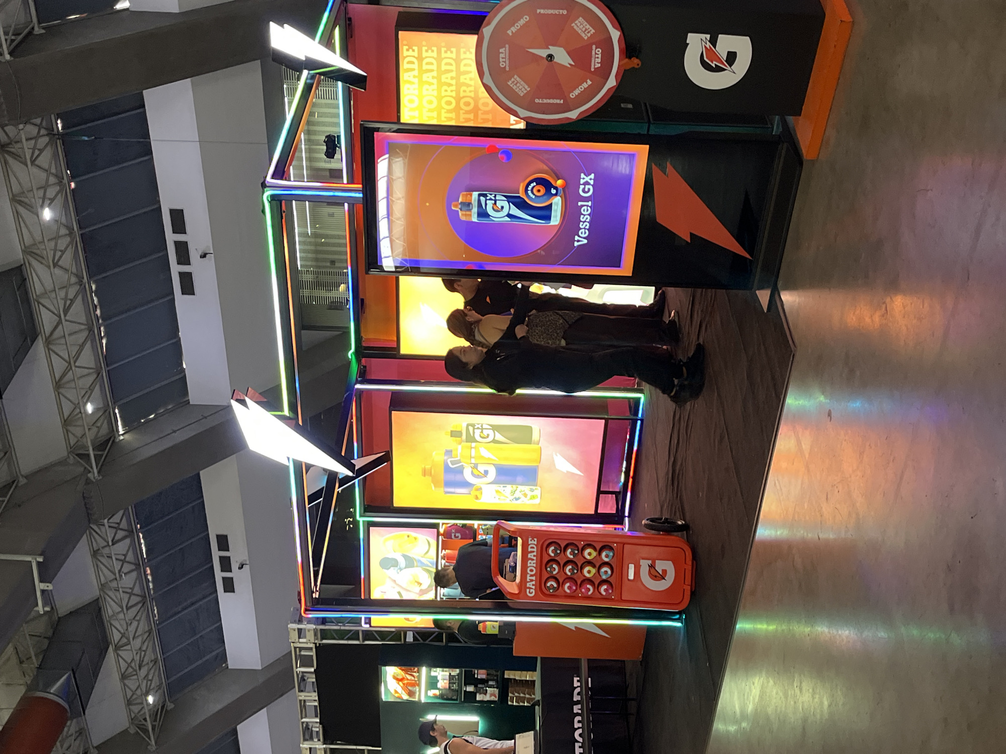 The image shows a promotional exhibit for Gatorade. The exhibit is located indoors, likely at a trade show or convention in the city of Puebla, Mexico (based on the "Puebla" in the filename). The Gatorade exhibit features several large video screens displaying animated product images with the text "Vessel GX", "TORADE", and "GATORADE". The booth has a brightly colored orange and black design, with neon lighting outlining the display. There is a large orange wheel with prizes written on it that are in Spanish. People are standing in front of the booth, interacting with the displays. There is a orange trolley-like car with many empty drink holders that has the Gatorade brand. A person is behind the trolley.