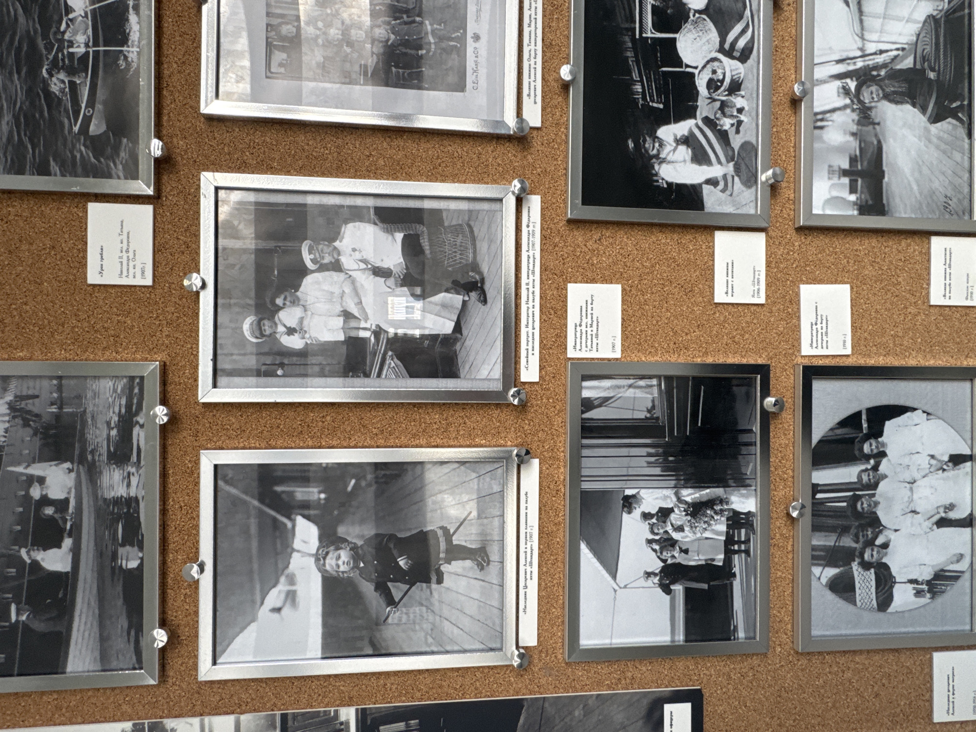 The image captures a corkboard displaying numerous framed, black and white photographs of the Russian Imperial Family. The photographs are held in place by metallic pushpins. The primary subjects include Emperor Nicholas II, Empress Alexandra Feodorovna, and their children: Grand Duchesses Olga, Tatiana, Maria, Anastasia, and Tsarevich Alexei.
The scene predominantly takes place aboard the Imperial yacht "Standart." Activities depicted include a "Rowing lesson" with Nicholas II, Tatiana, Alexandra Feodorovna, and Olga (1907), family portraits on the yacht's deck, and candid shots of the children. Tsarevich Alexei is notably featured in a photograph from his "first voyage" on the "Standart" (1907). Other photos show groups of the Grand Duchesses and Tsarevich, as well as Empress Alexandra with her daughters.
Notable details include the period attire of the subjects, often naval-style uniforms for the men and boys, and elegant dresses for the women and girls. The photographs are dated from 1906 to 1910, offering a glimpse into the family's life during this era. The frames are thin and metallic, casting slight reflections. The corkboard background provides a textured contrast to the smooth, glossy surfaces of the framed images. The photographs are arranged in an informal, overlapping grid.