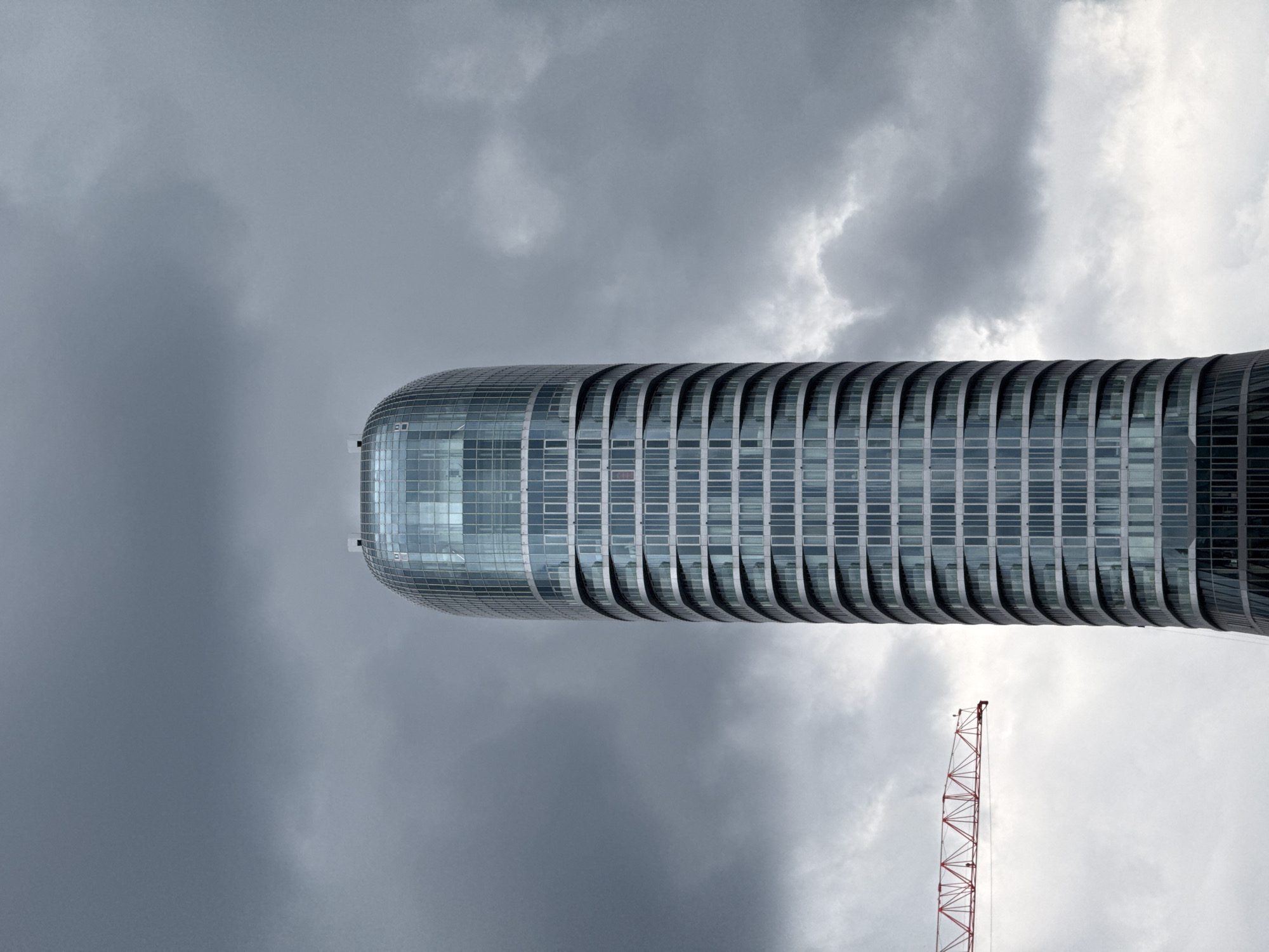 The image displays a tall, modern skyscraper set against a dramatic, overcast sky. The building, which appears vertically oriented in its intended perspective, showcases a distinctive curvilinear architectural design, possibly cylindrical or oval. Its facade is predominantly glass, featuring a grid of numerous rectangular windows. A prominent architectural detail is the series of horizontal, projecting bands or fins that encircle the building, giving it a ribbed or textured appearance along its length. The top of the skyscraper is smoothly rounded.
The sky is largely covered by heavy, grey clouds, with some brighter patches indicating varying cloud thickness. In the lower right corner of the frame, a section of a red and white construction crane is partially visible, suggesting ongoing development in the urban environment.
The scene depicts a contemporary urban landscape during daytime, under cloudy weather conditions. No specific activity or individuals are directly observed. The modern architecture of the skyscraper and the presence of a crane are characteristic of a bustling city. This architectural landmark is situated in Belgrade, Serbia. There is no visible text in the image.