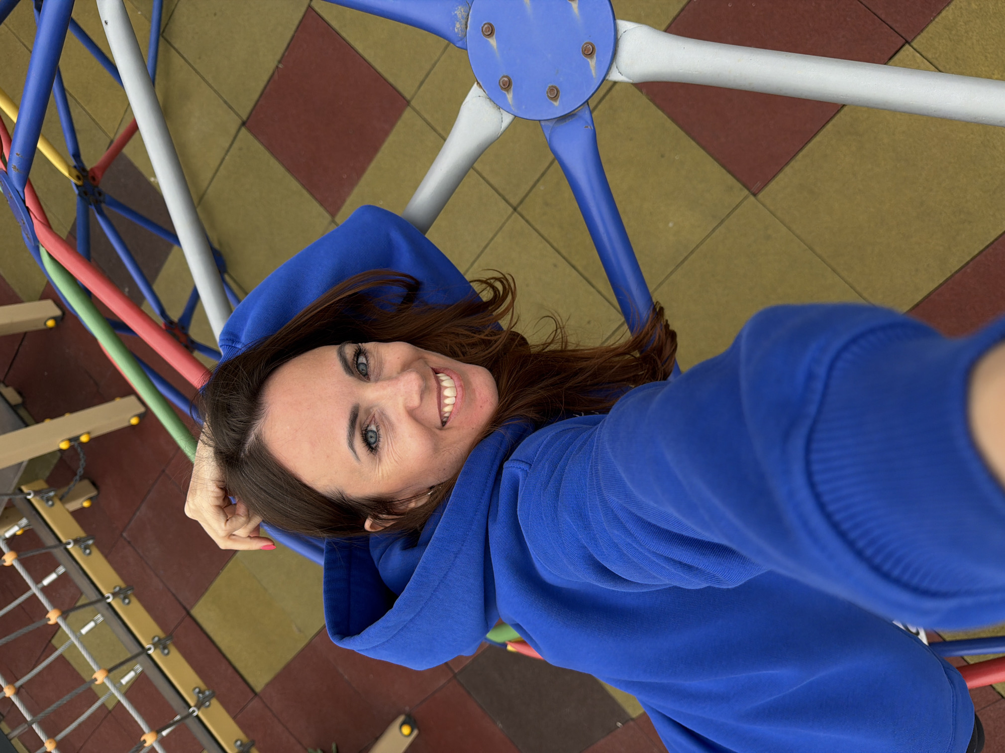 A woman is lying on a playground structure, taking a selfie. She has brown hair, blue eyes, and fair skin, and she is wearing a blue hoodie. The playground structure consists of blue, white, red, green, and yellow bars. The ground underneath the playground structure has a tile pattern of red and yellow squares. The location is Birobidzhan, Russia.
