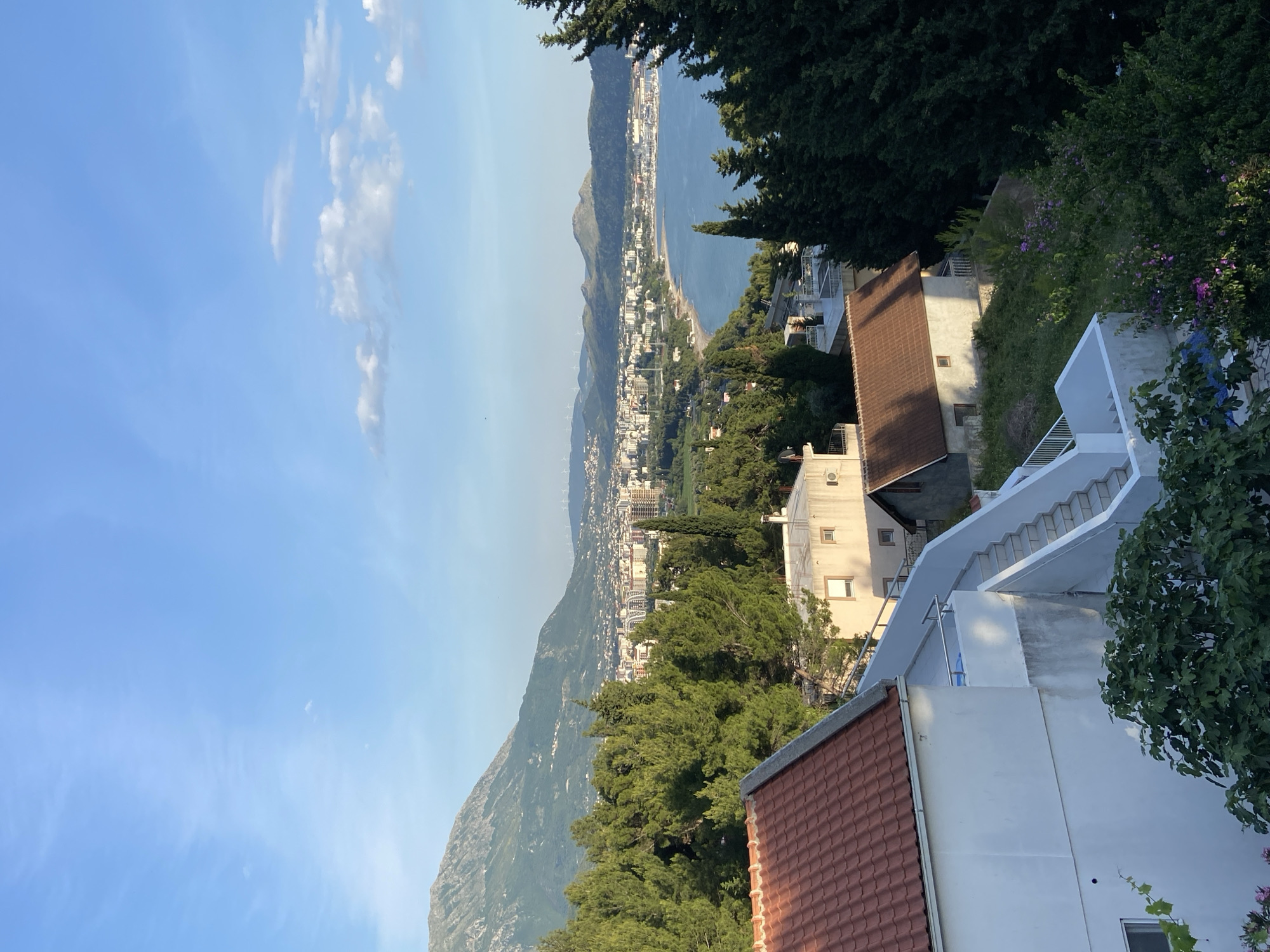 The image depicts a view of Šušanj, Montenegro, from an elevated position. In the foreground, there are rooftops of buildings, some with red tiles and others painted white. White staircases are visible leading up to higher levels. Lush green trees and vegetation surround the buildings. In the midground, a densely populated town stretches along the coastline. The town is nestled between the Adriatic Sea and a rugged mountain range. The sea is calm and blue. The sky is clear with scattered clouds. The overall scene is bathed in sunlight, suggesting it is daytime.