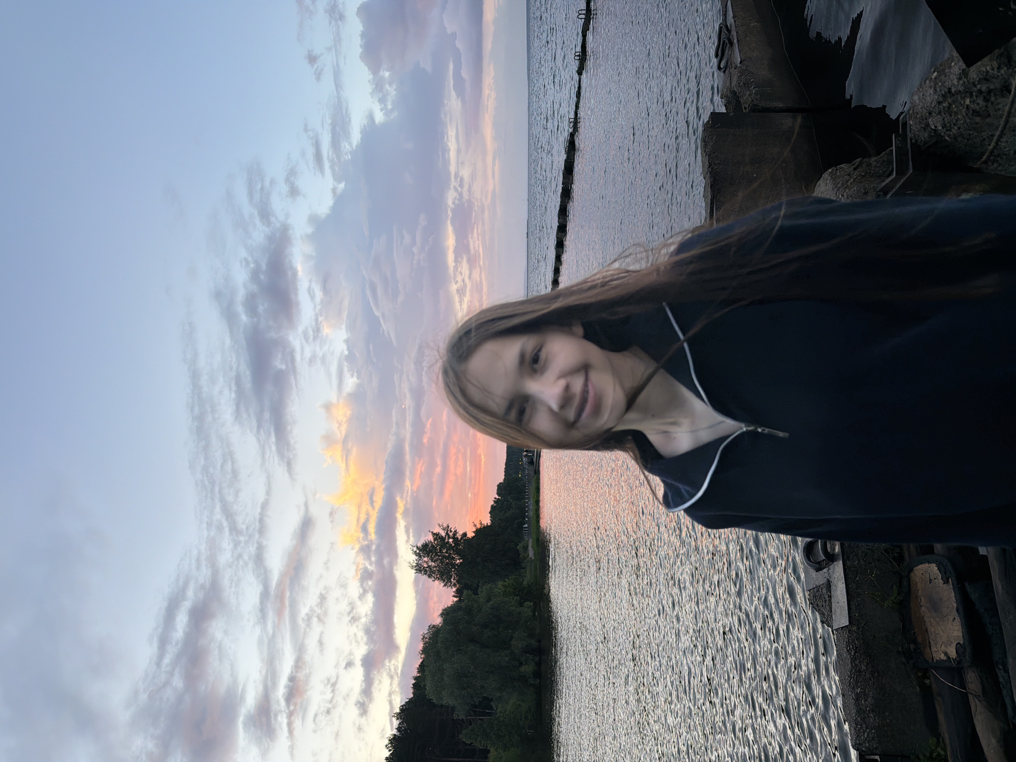 The image depicts a young woman standing on a concrete pier by a body of water, with a forest and a pink and orange sunset sky in the background. The woman is facing forward and smiling. She is wearing a dark blue zippered hoodie with white trim. The pier is made of rough concrete blocks and has some dark debris in the foreground. The water is calm with ripples and reflects the colors of the sunset. The sky has scattered clouds that are illuminated by the setting sun. The location appears to be a waterfront area, possibly a park or recreational spot, in the city of Chaikovsky, Russia, given the context provided.