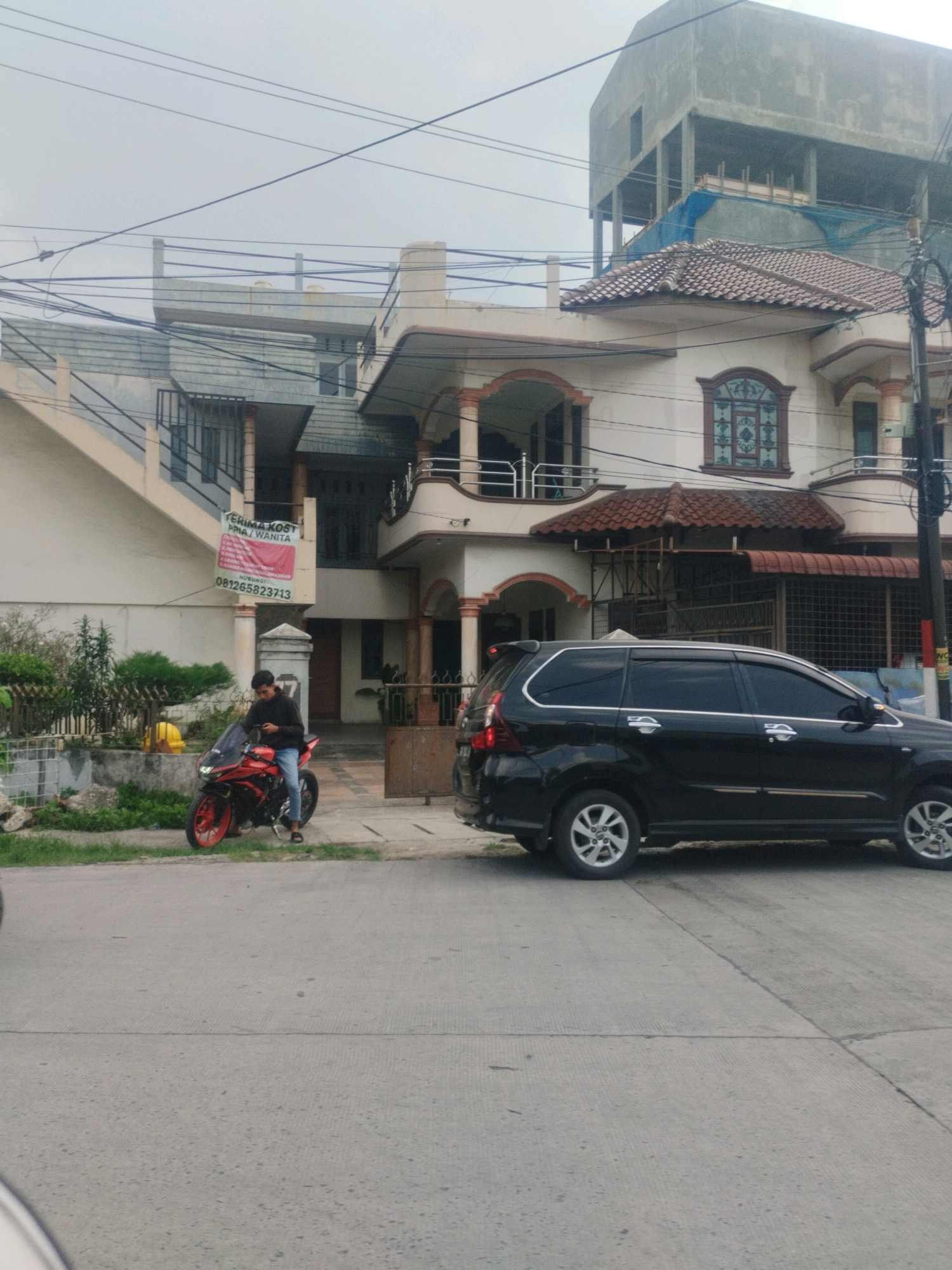 A man in a black long-sleeved shirt and blue jeans is sitting on a red motorcycle parked on the side of a street in the city of Medan, Indonesia. The man is looking down at his phone. To the right of the motorcycle, a black SUV is parked. In the background, there are multi-story buildings. One building has a large sign that says "TERIMA KOST PRIA/WANITA" which translates to "GUEST HOUSE FOR MEN/WOMEN." This indicates the area is residential and likely a commercial area for rental properties. The sky is overcast.
