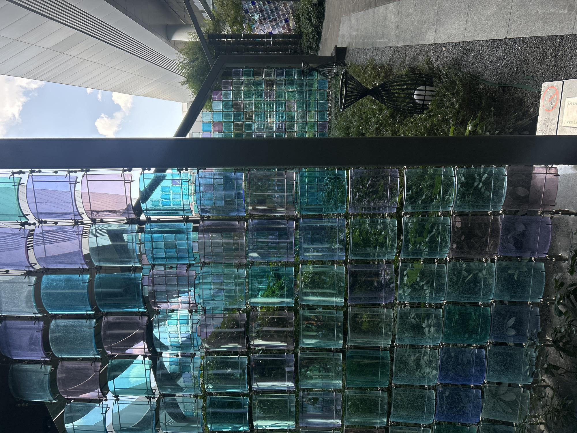 In Jianyang City, China, a modern architectural design features a striking vertical installation composed of numerous translucent, cylindrical glass elements. These glass pieces are arranged in a grid-like pattern and display a gradient of colors ranging from deep blues and greens to purples and lighter blues, with some appearing almost clear. The glass cylinders are segmented and held together by a dark, metallic framework, creating a layered, textured effect. Within many of these glass units, lush green foliage is visible, suggesting the installation may serve as a vertical garden or display.
The installation stands adjacent to a paved walkway and is surrounded by dense, verdant shrubbery. Two ornate, black metal sculptures, resembling hourglasses with a spherical object inside each, are placed within the greenery. Beyond the walkway, a modern building with a sleek, gray facade and angular architectural lines dominates the background. Sunlight illuminates the scene, casting shadows and highlighting the translucency and colors of the glass installation. The sky above is a clear blue, dotted with a few white, puffy clouds. The overall impression is one of contemporary urban design integrating natural elements through an artistic, colorful structure.