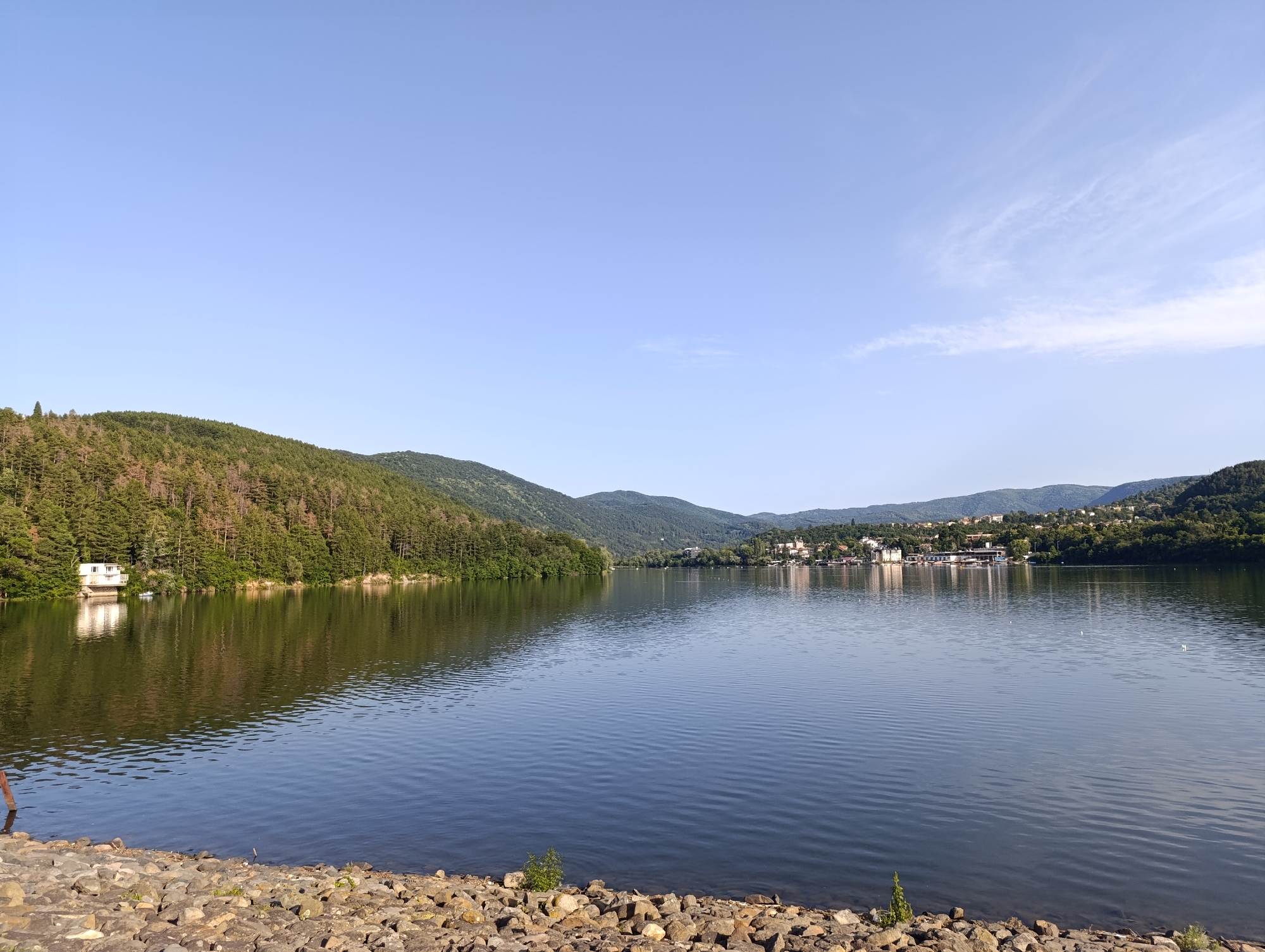The image displays a wide, calm lake reflecting the surrounding lush green hills under a clear blue sky. In the foreground, a rocky shoreline is visible. On the left side of the lake, a white building with a red roof sits on the edge of a densely wooded hill. Across the water, a town with various buildings and structures is nestled amongst the trees on a hillside, with some boats docked along the shore. The scene suggests a peaceful, natural environment within or near an urban area. This specific location is identified as Pancharevo Lake, located in Pancharevo, Bulgaria.