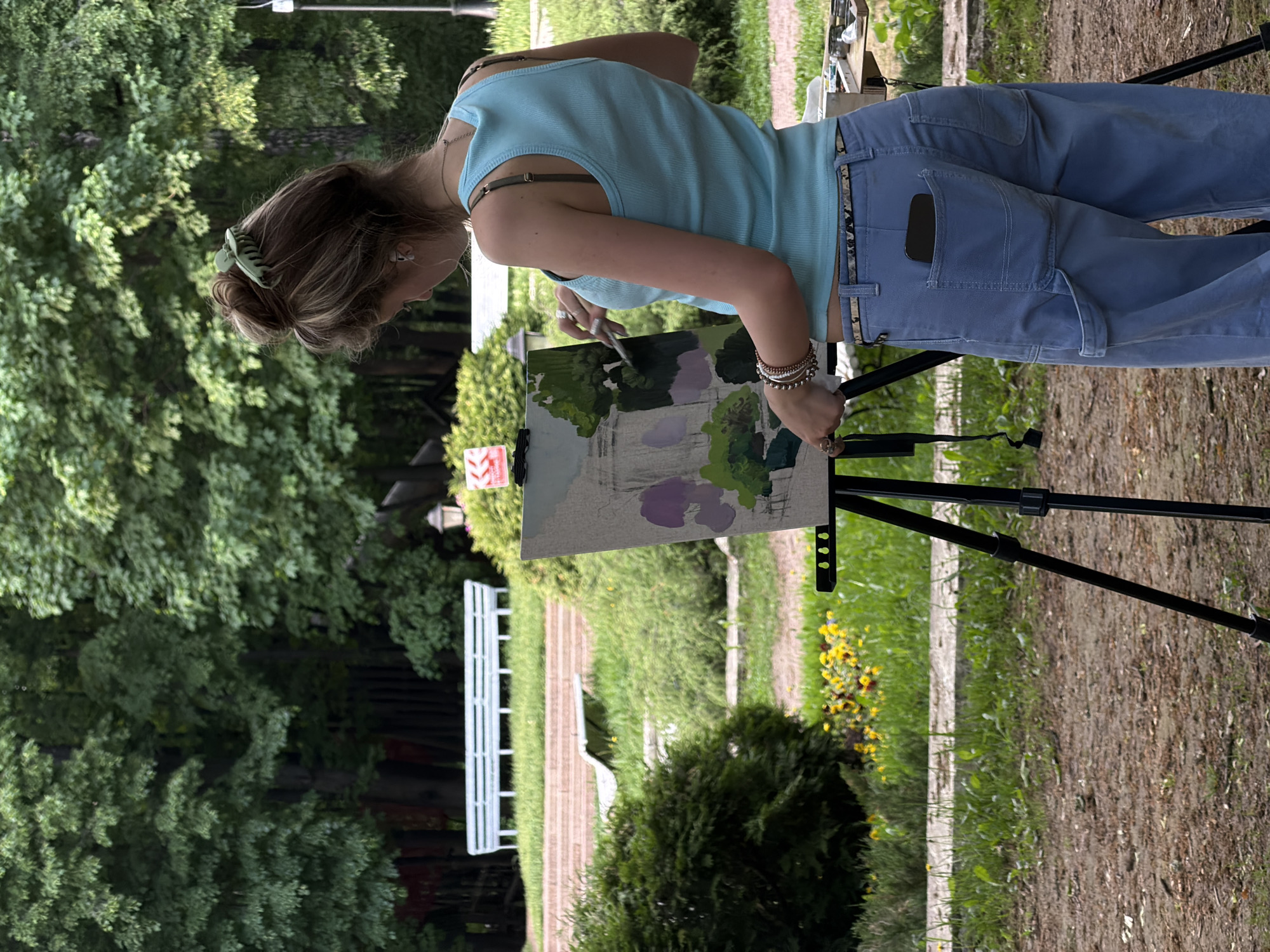 A woman is painting outdoors in a park, likely in Moscow, Russia. She is fair-skinned with light brown hair pulled back with a green clip. She is wearing a light blue tank top and light blue cargo pants with a patterned belt. Her phone is visible in her back pocket. She is standing at an easel, painting a landscape. The painting includes trees, grass, and possibly a structure with columns, painted with green, purple, and grey hues. The park has benches, grass, trees, and a stone path. Yellow flowers are growing near the path. The ground is dirt and grass. The image is oriented vertically.