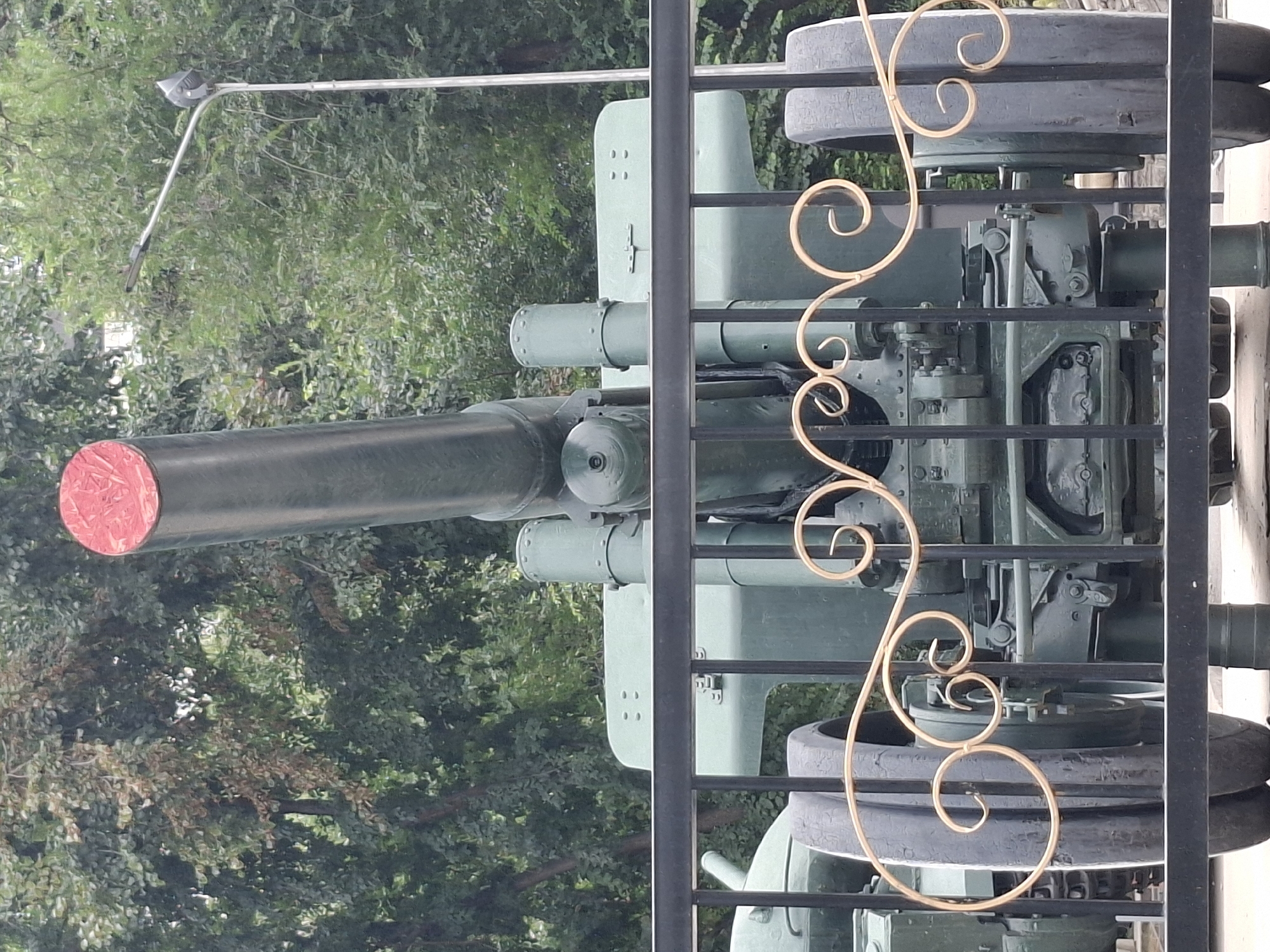 The image shows a large, dark green artillery gun, likely a historical monument, positioned behind a black metal fence with ornate gold scrollwork. The gun's barrel, which is quite prominent and appears to be capped with a red material, points upwards. The weapon is mounted on a wheeled carriage, with two large tires visible on each side. The background consists of dense green foliage, suggesting an outdoor setting with trees. A tall, silver lamp post is visible to the right of the gun.
While the image does not provide explicit visual cues to confirm its location within Almaty, Kazakhstan, this particular artillery piece is a well-known monument located in the Park of 28 Panfilov Guardsmen. This park is a significant landmark in Almaty and houses various war memorials. The artillery piece, specifically a 1944 M-30 122 mm howitzer, is part of the memorial complex dedicated to World War II. The presence of such a monument is consistent with its placement in a prominent city park in Almaty, Kazakhstan.