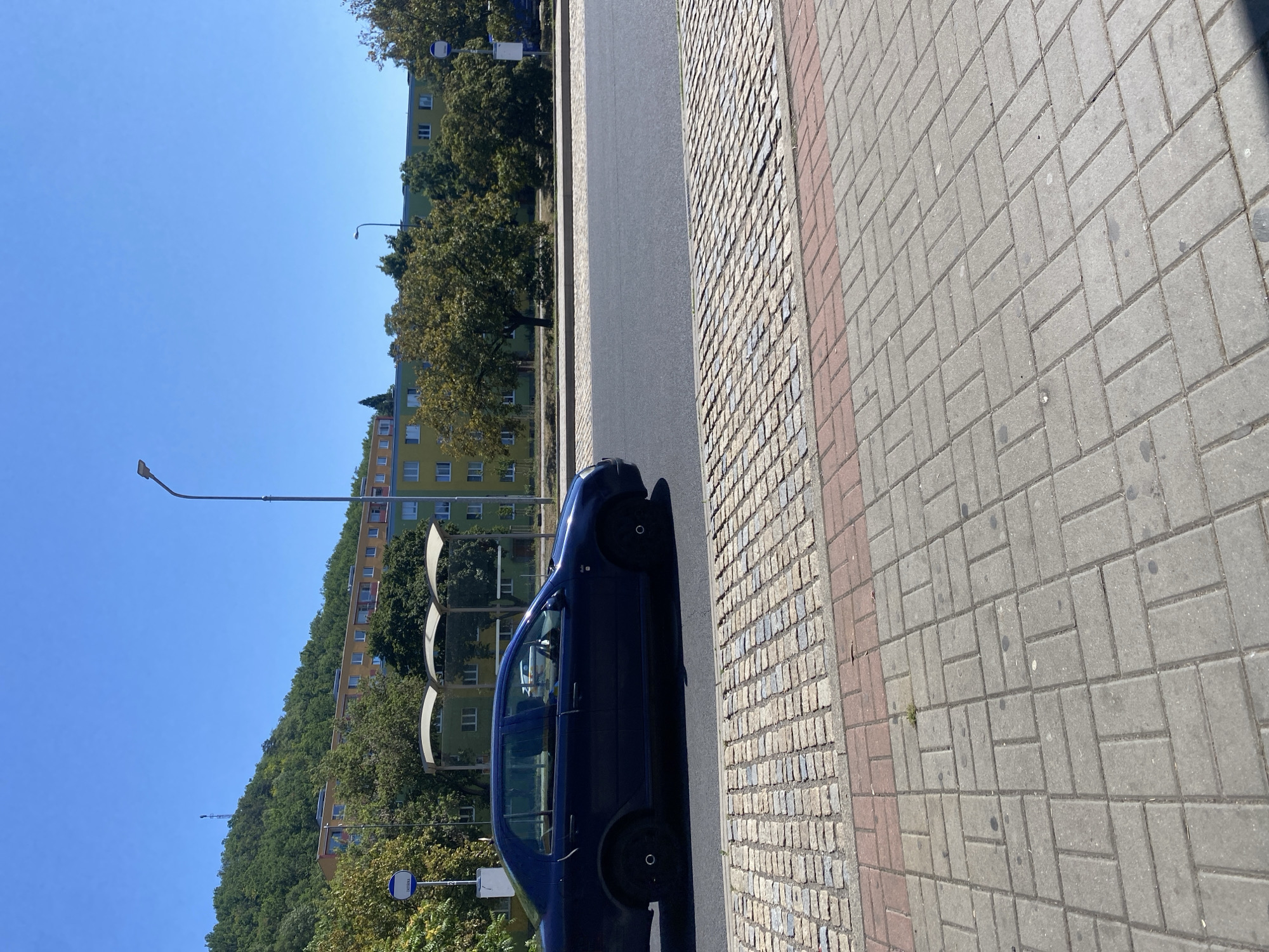 A dark blue sedan is driving on a street in Bílina, Czechia, on a clear, sunny day. The car is in motion, and its side is visible from the left. The street is lined with trees and a sidewalk made of grey and red paving stones. In the background, there is a hill covered with lush green trees, a multi-story building with yellow and green facades, and a bus stop. A streetlamp is visible on the right side of the image. The sky is a vibrant, cloudless blue.
