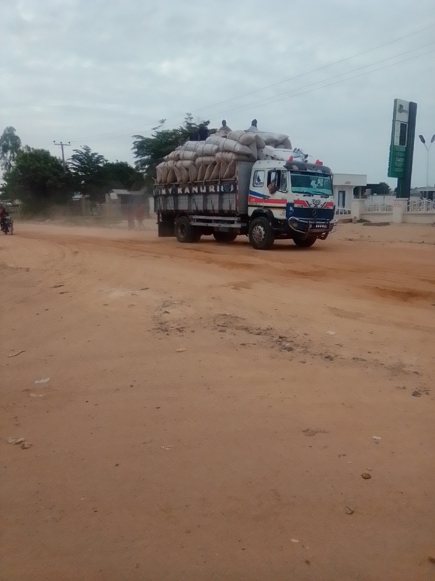 A wide-angle, eye-level, slightly dusty, outdoor shot captures a truck loaded with sacks on a dirt road in Gombe, Nigeria. The truck, a white Mercedes-Benz model, is traveling from left to right and is heavily laden with numerous light-colored sacks, some secured with netting. Two men are visible on top of the sacks, appearing to be workers. The truck has red and blue accents on its cab, and the text "M. KANURIO" is visible on the side of the trailer. A few other people and a motorcycle are also present further down the road, obscured by dust kicked up by the truck. In the background, there are trees, a light pole, a white building, and a tall, green signpost with white lettering, possibly indicating a fuel station. The sky is overcast with a pale, diffused light. The road is unpaved and appears to be made of reddish-brown earth.