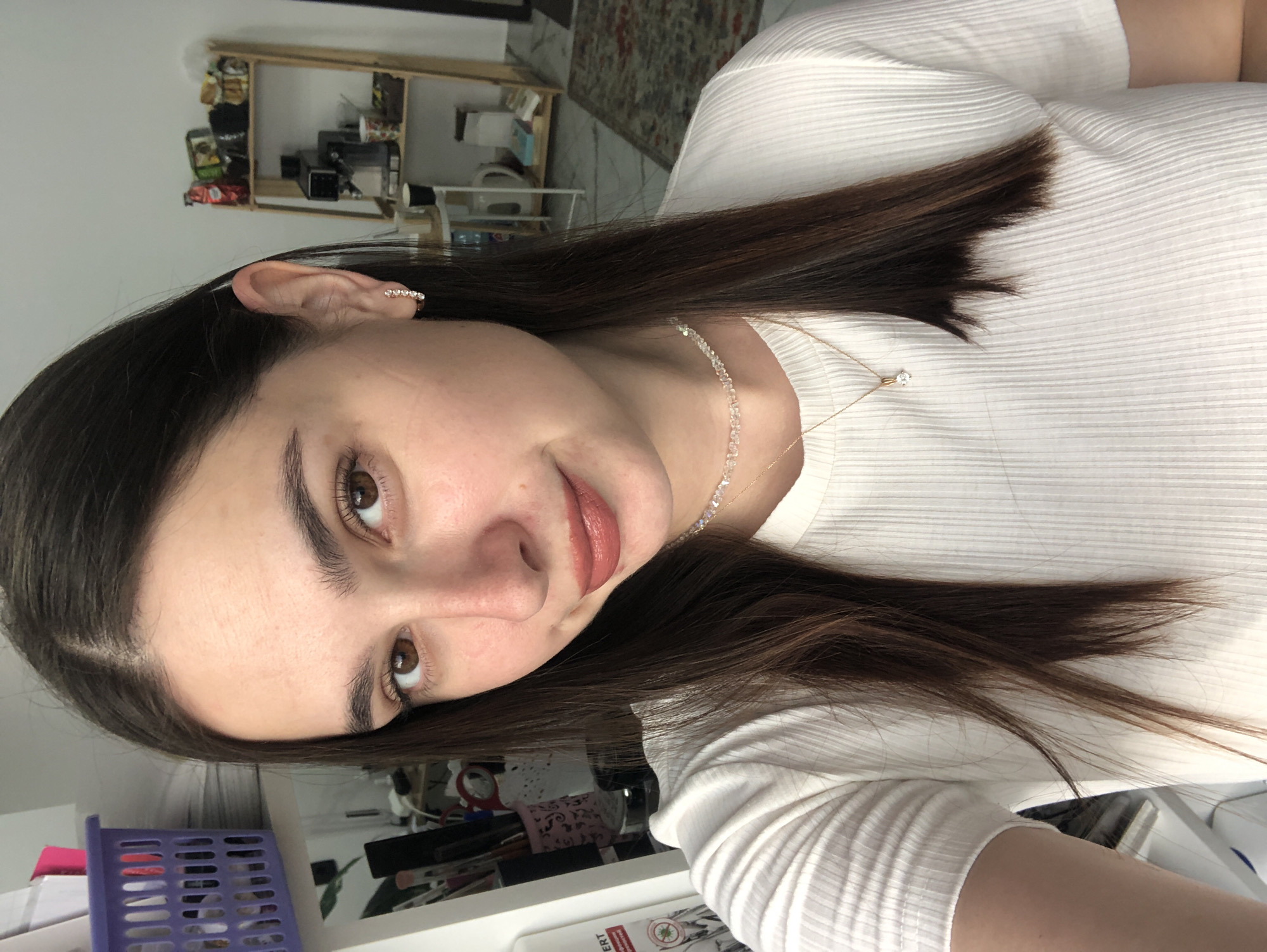 A young woman with long, dark brown hair and brown eyes is taking a selfie in a room. She is wearing a white, ribbed t-shirt and a gold necklace with a diamond pendant, and a small diamond earring in her left ear. Her lips are painted with a reddish-brown lipstick. In the background, there are shelves with various items, including a coffee maker, cups, and boxes. To the left of the woman, there is a purple laundry basket and some paperwork. The floor has a patterned rug. The scene appears to be an indoor domestic environment, possibly a home or apartment, and there is no visible indication of the city of Chaikovsky, Russia.