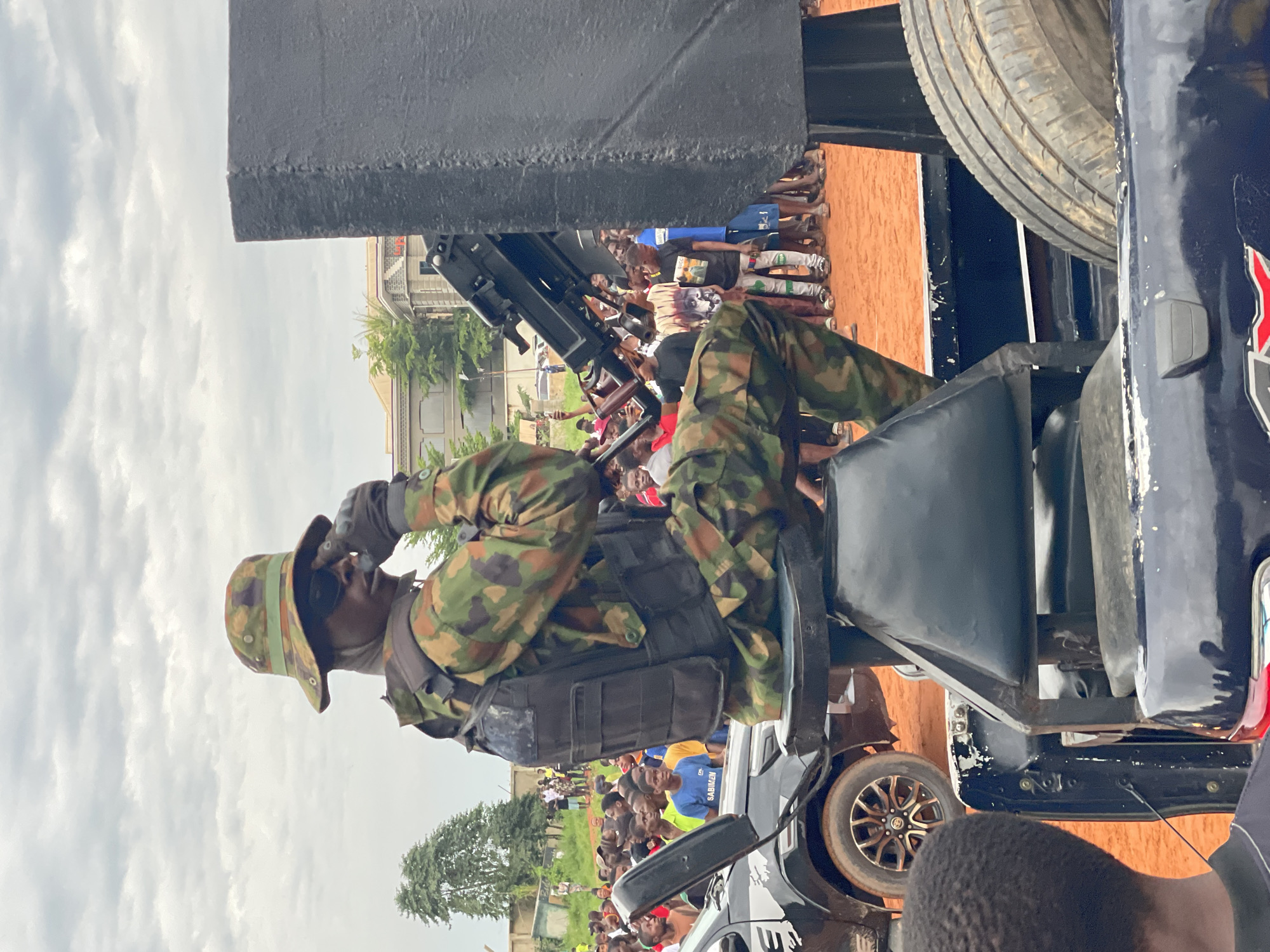 A soldier in camouflage fatigues and a bucket hat sits on a vehicle, holding a firearm. The soldier is wearing sunglasses and gloves. A crowd of onlookers, mostly men and boys, are gathered behind the vehicle. The ground is covered in reddish-brown dirt, and a few buildings and trees are visible in the background. The sky is overcast with clouds. The overall impression is of an outdoor event or demonstration, possibly a public appearance by the military.