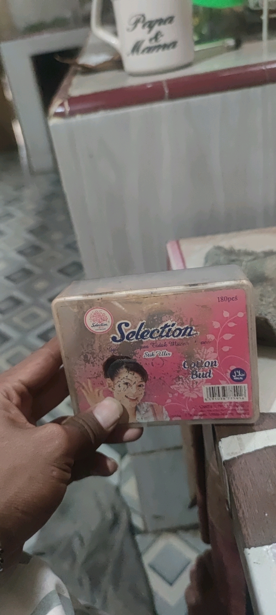 The image shows a person's hand holding a package of "Selection" brand cotton buds. The package is pink and features an image of a young woman. Text on the package includes "180pcs", "Cotton Bud", and "HALAL". The background appears to be indoors, with tiled flooring visible. The mug in the background has "Papa & Mama" written on it. The context of Gambarsari, Indonesia, is not directly evident from the visual information in the image.