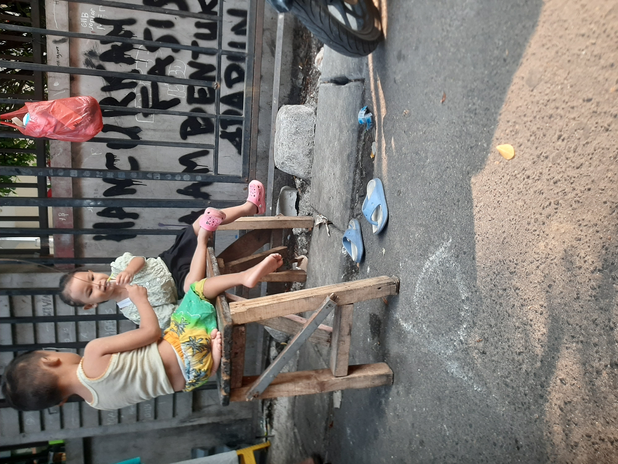 In Central Jakarta, Indonesia, two young children are seated on a makeshift wooden stool. The child on the left, wearing a white tank top and brightly colored shorts with a palm tree pattern, faces away from the camera, with their legs dangling. The child on the right, wearing a pale green shirt and a black skirt, faces the camera and appears to be holding a thin twig or piece of grass to their mouth. A red plastic bag hangs on a metal fence behind them, which is covered in graffiti. The ground is paved with asphalt and concrete, with scattered debris including a blue pair of flip-flops. A motorcycle wheel is visible on the right side of the frame.
