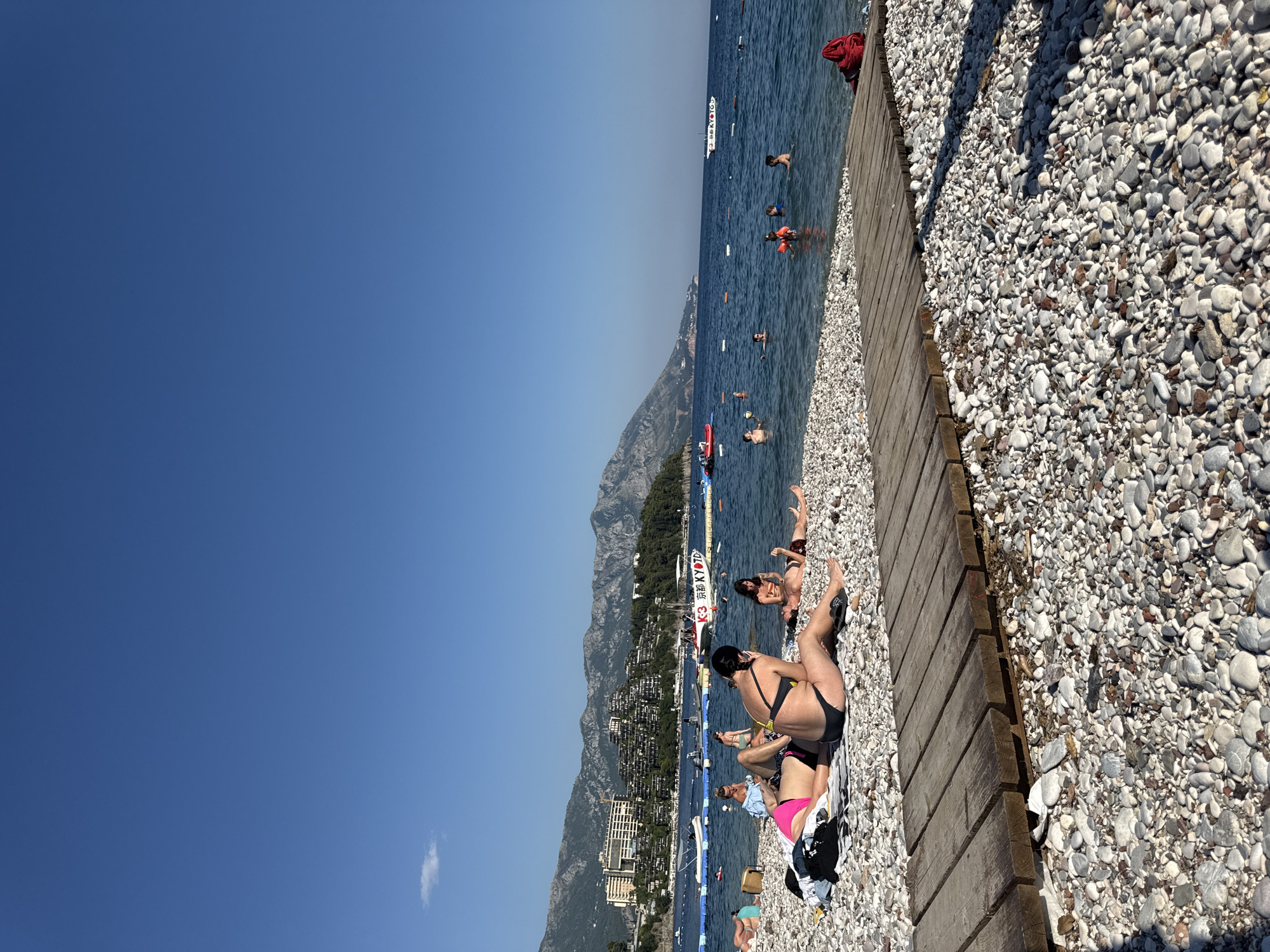 This is a photograph of Mogren Beach in Budva, Montenegro, on a clear, sunny day. The beach is composed of pebbles, with a wooden walkway in the foreground. Several people are visible on the beach, sunbathing and relaxing. Some are wearing swimwear. In the sea, there are people swimming and enjoying the water. Various boats and a floating platform are also present in the sea. In the background, mountains covered with buildings and lush vegetation rise up, forming a scenic backdrop to the beach. The sky is a vibrant blue, with a single small cloud.