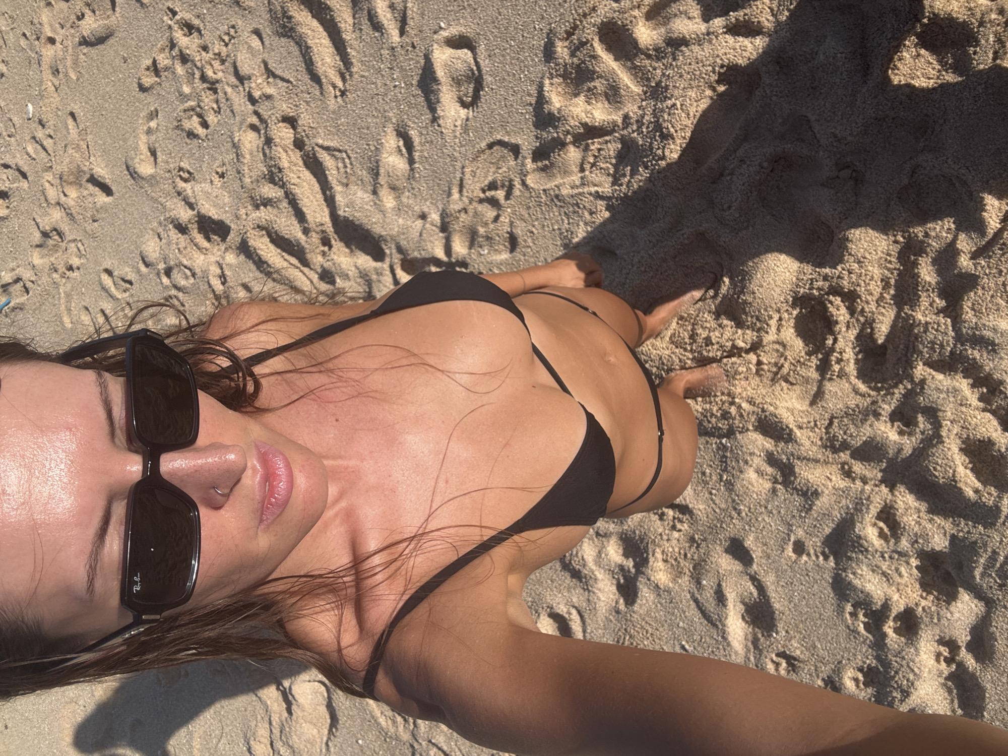 A woman wearing a black bikini and sunglasses is taking a selfie on a sandy beach. She is standing on the sand, which is imprinted with footprints and textured by the sunlight. The woman is looking down at the camera and her body is angled towards it. The background is a blurred expanse of sand and the ocean, suggesting a tropical location like Kuta Selatan, Indonesia. The lighting indicates it is daytime, likely late afternoon due to the long shadows cast on the sand.