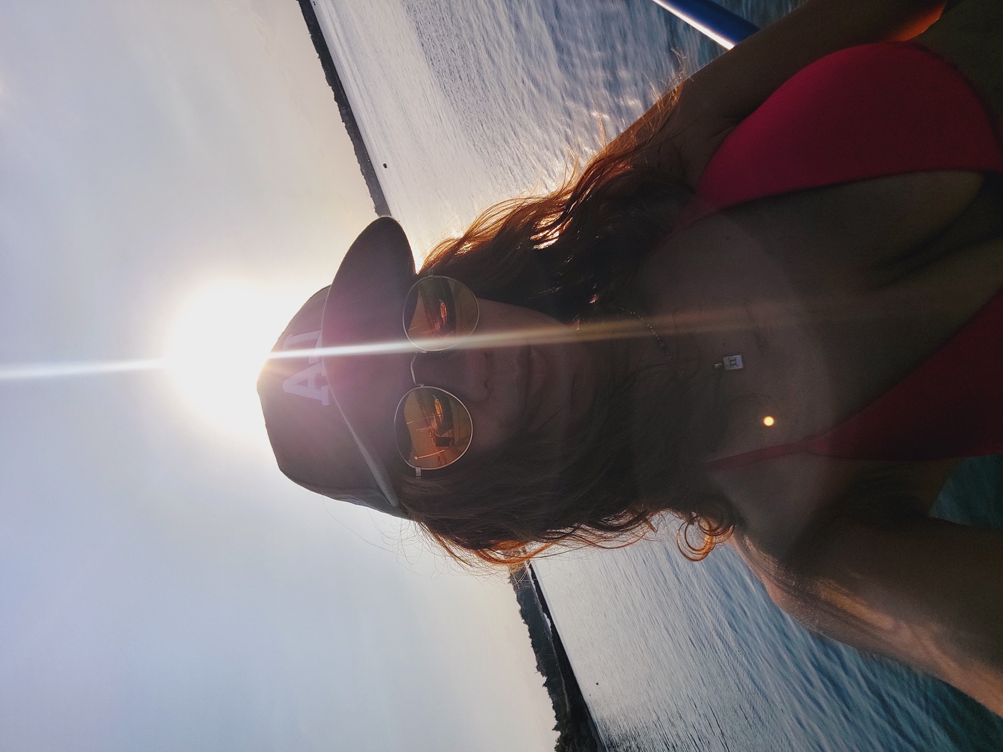 A woman wearing a red bikini, sunglasses, and a baseball cap is captured in a selfie taken outdoors with a body of water and a hazy sky. The sun is positioned behind her, causing a lens flare. The water appears calm, with a distant shoreline visible. The woman has long, reddish-brown hair. The scene is consistent with an outdoor recreational setting, possibly near the coast, in a location like Nesebar, Bulgaria, known for its historical sites and Black Sea coast.