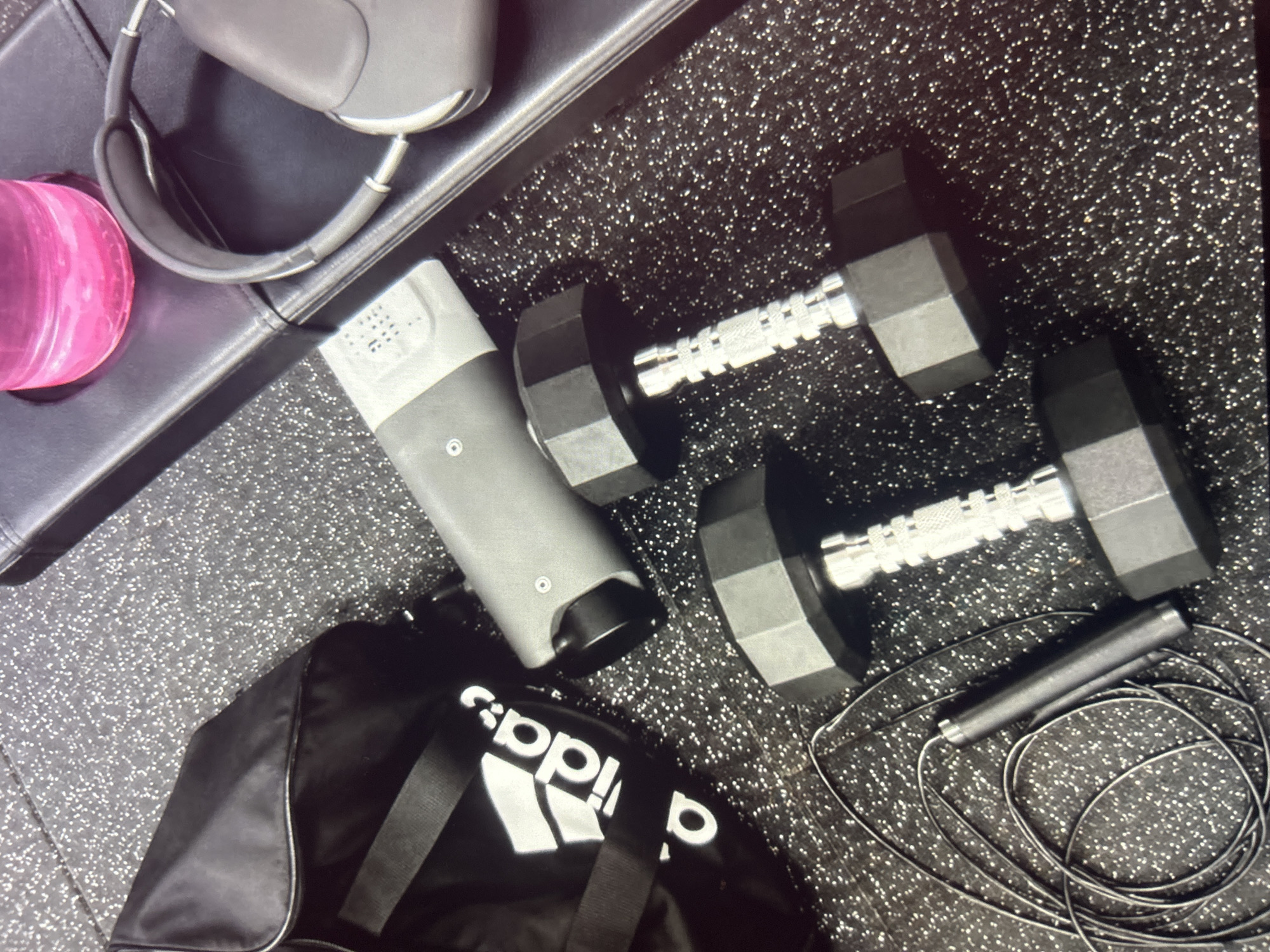 This is a medium-shot, high-angle, eye-level, indoor, close-up, photographic still of a fitness equipment arrangement. The scene appears to be set on a black, speckled gym floor.
Prominently featured are two hexagonal dumbbells with chrome handles. To the left of the dumbbells, a black Adidas gym bag is visible, adorned with the white Adidas logo. A black jump rope is coiled on the floor in the lower left corner.
In the upper right quadrant of the image, a black padded bench or surface holds a pink liquid in a clear bottle and a pair of gray over-ear headphones. A gray cylindrical object with some textured detailing and a circular indentation is positioned between the gym bag and the dumbbells. The overall impression is one of preparedness for a workout. The image's perspective and subject matter suggest it was taken in a personal or commercial gym environment, possibly in Knezha, Bulgaria, given the prompt.