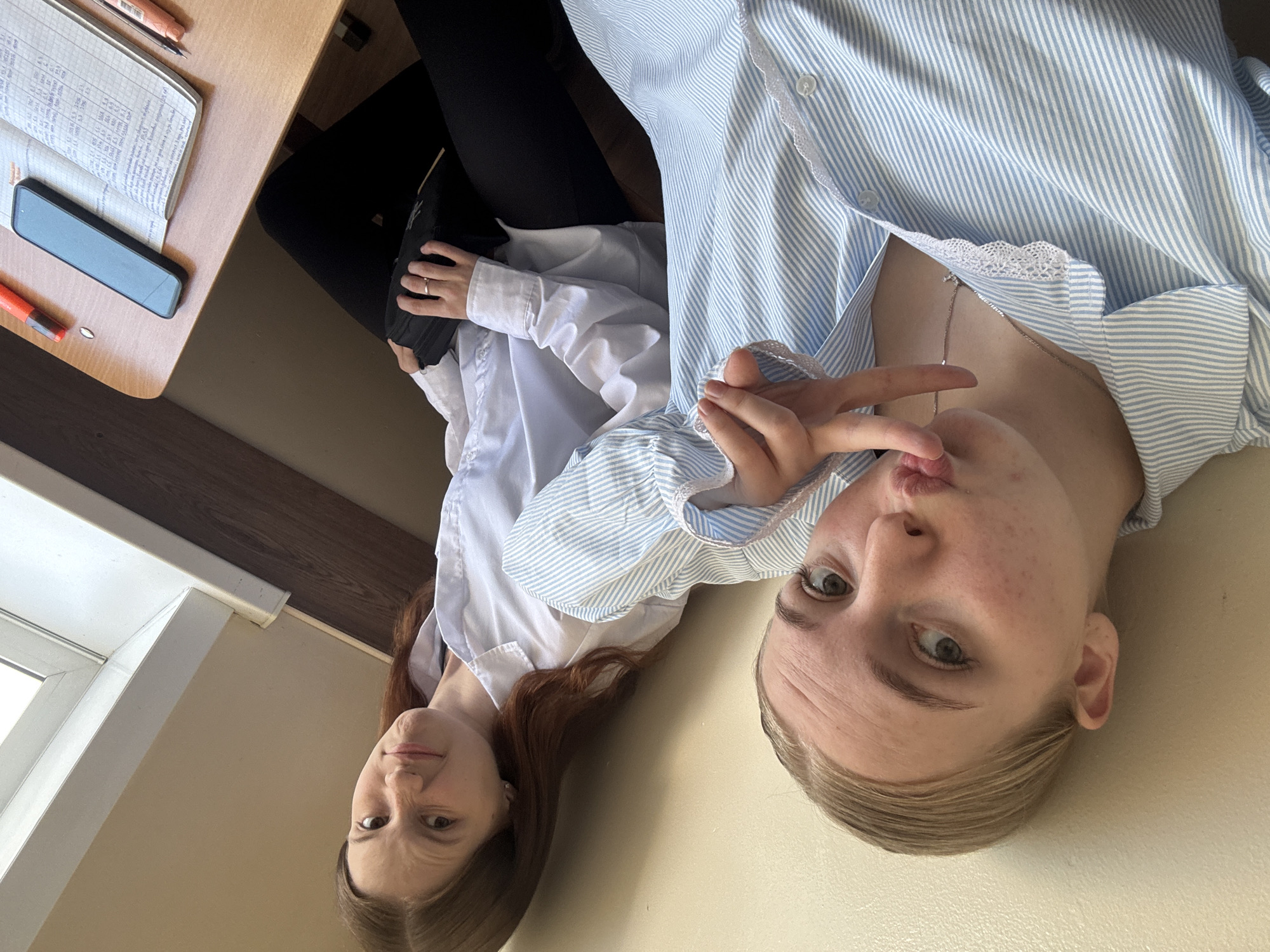 Two young women pose for a selfie indoors. One woman is on the left with shoulder-length brown hair. She wears a white button-down shirt. The other woman is on the right with blonde hair. She wears a striped blue and white button-down shirt with lace trim. Her hands are in a peace sign, posing near her face. They sit at a desk with a notebook and a phone on it. The location may be Perm, Russia.