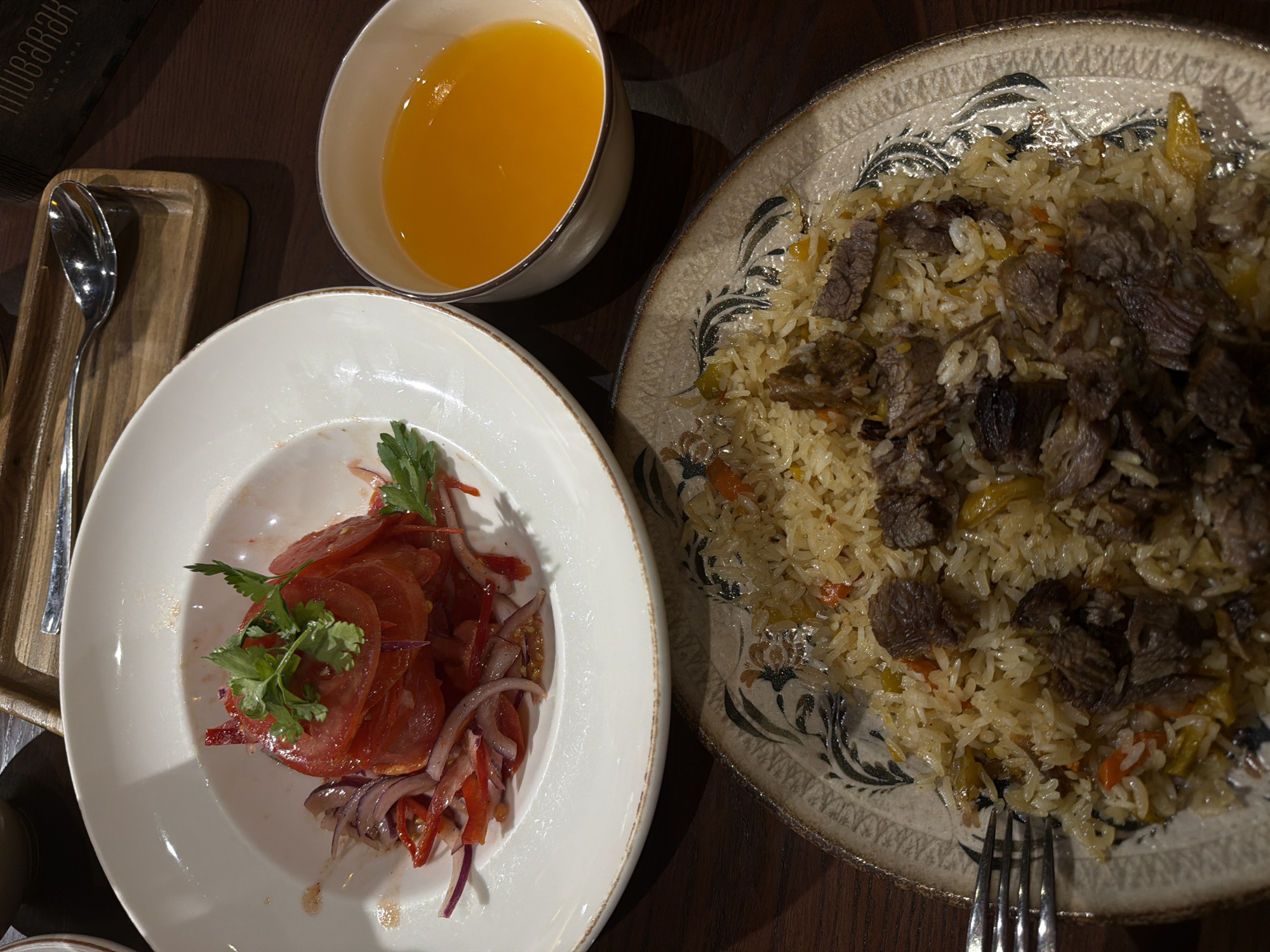 A still life of food, likely from a restaurant, is depicted from a top-down perspective. In the foreground, a plate of pilaf, a rice dish with meat and vegetables, is prominently displayed. To the left, a white bowl holds a sliced tomato salad with onions and herbs. Adjacent to the salad, a small bowl contains a bright orange liquid, likely juice. In the background, a wooden tray with a spoon rests on a dark wooden table. A portion of a box with Arabic script is visible in the upper right corner.