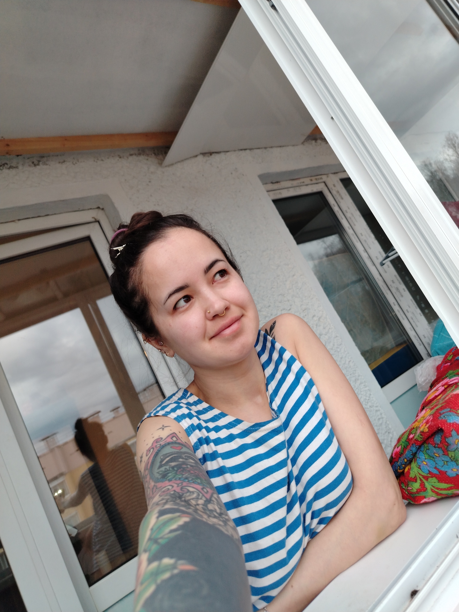 A woman sits in front of a window, wearing a blue and white striped tank top. She has dark hair pulled back in a bun, with piercings in her nose and ears. Her left arm, which is heavily tattooed, is visible in the foreground. The woman is looking slightly upwards and to the right, with a subtle smile. The window frame is white. Reflected in the window is a silhouette of a figure and nearby buildings. The background outside the window appears to be a cloudy sky and distant trees. The building appears to be a light colored concrete structure. A colorful blanket is next to her on the window sill.
Given the information, "location in the city of Noviy, Russia," this could be interpreted as information about the location, but it's not verifiable from the image alone.