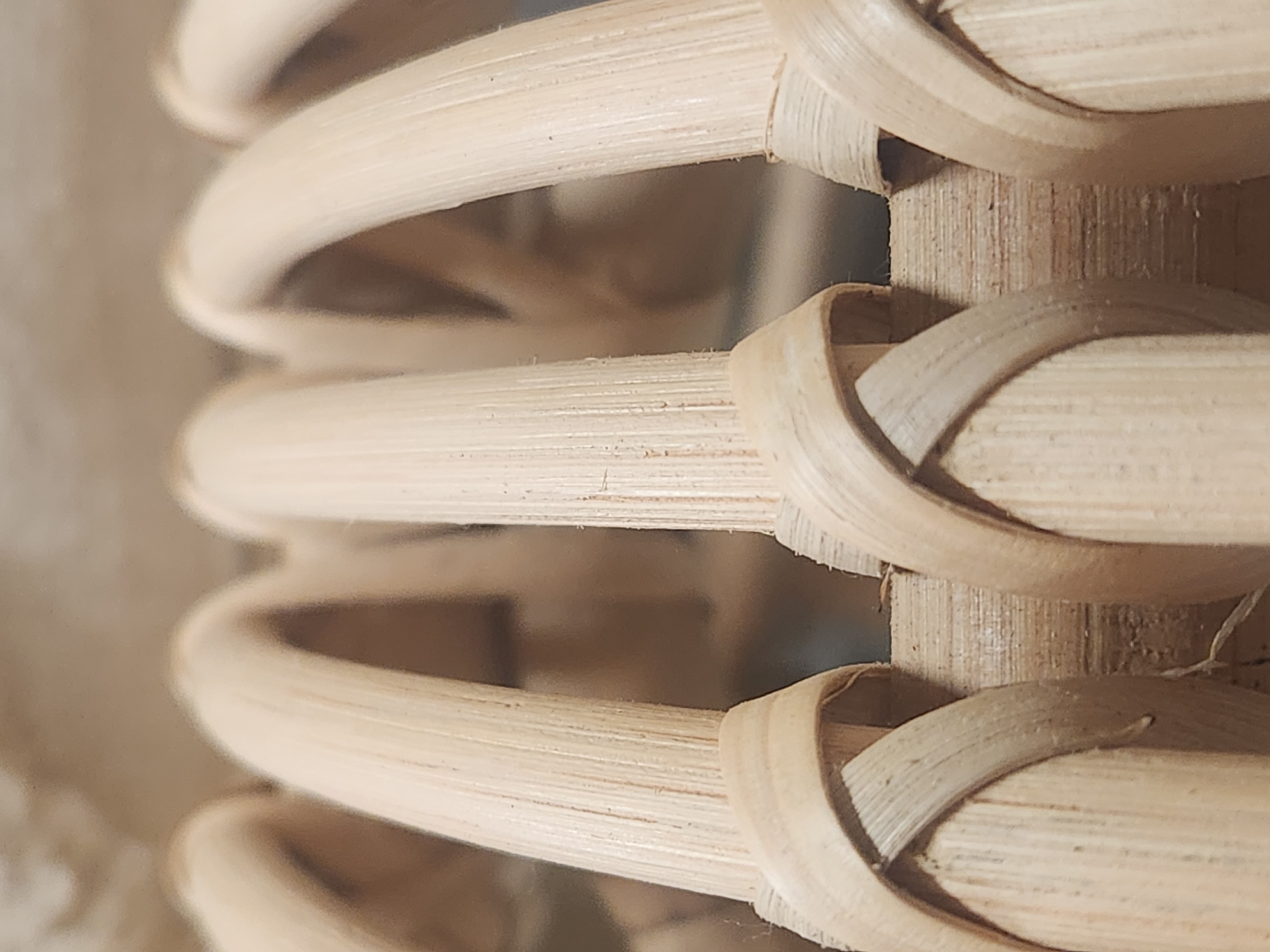 The image shows a close-up, eye-level shot of woven bamboo or rattan material, likely part of a piece of furniture. The material is light brown and appears to be made of thin, hollow reeds. The weaving pattern consists of horizontal reeds interlaced with vertical strands. The texture is rough and fibrous. The background is blurred and appears to be a neutral, muted color. There are no people, animals, or any other discernible objects present in the scene. The image provides no information about its location in Maitland, United States of America, or any specific context regarding its origin or purpose.