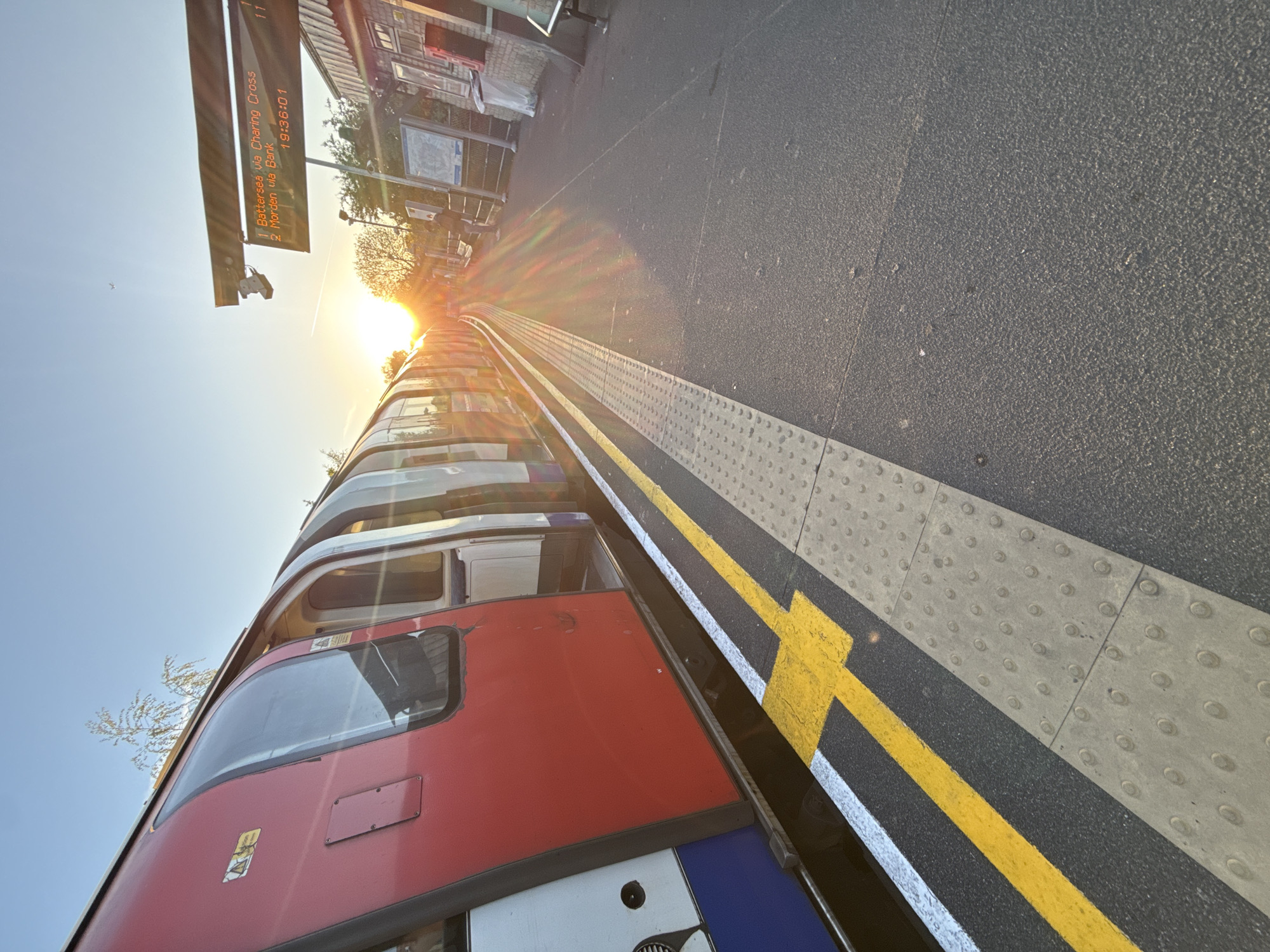 The image depicts a train platform in London, United Kingdom, at sunset. A London Underground train, painted in red, white, and blue, is situated alongside the platform. A yellow line is painted on the edge of the platform to denote the safe area for passengers. Above the train is a display board showing train arrival information: Battersea via Charing Cross and Morden via Bank, with the time 19:36:01. The sun is setting at the end of the train tracks.