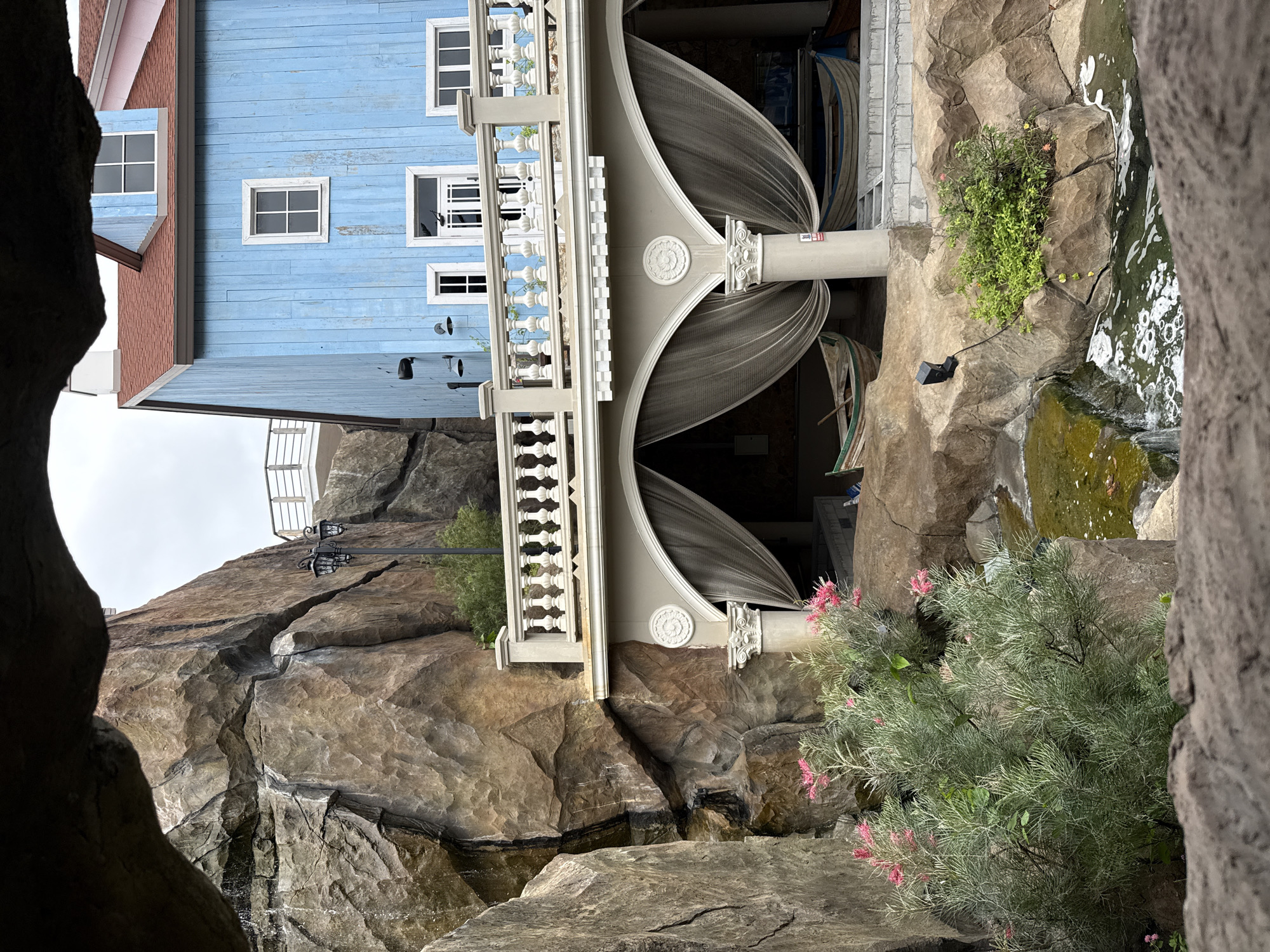 The image depicts an outdoor scene, likely in a tourist area, with a focus on artificial rock formations and a blue wooden building. The perspective is from within a cave-like opening, looking out. To the left, a large, textured rock formation dominates the frame, with some greenery and pink flowers in the foreground. Water appears to be trickling down the rock face in places.
In the center and right of the image, a two-story blue wooden building with a red roof is visible. It has white-framed windows and doors. A white balcony with a balustrade extends from the upper floor. Below the balcony, an open-air area is draped with sheer curtains, and several boats are parked or displayed within this sheltered space. The ground is covered with more artificial rock formations and a small body of water with some ripples. The sky is overcast. The style of the architecture and landscaping suggests a themed attraction or resort. The image does not contain any people. There is no information in the image itself to identify the location as Pattaya City, Thailand.