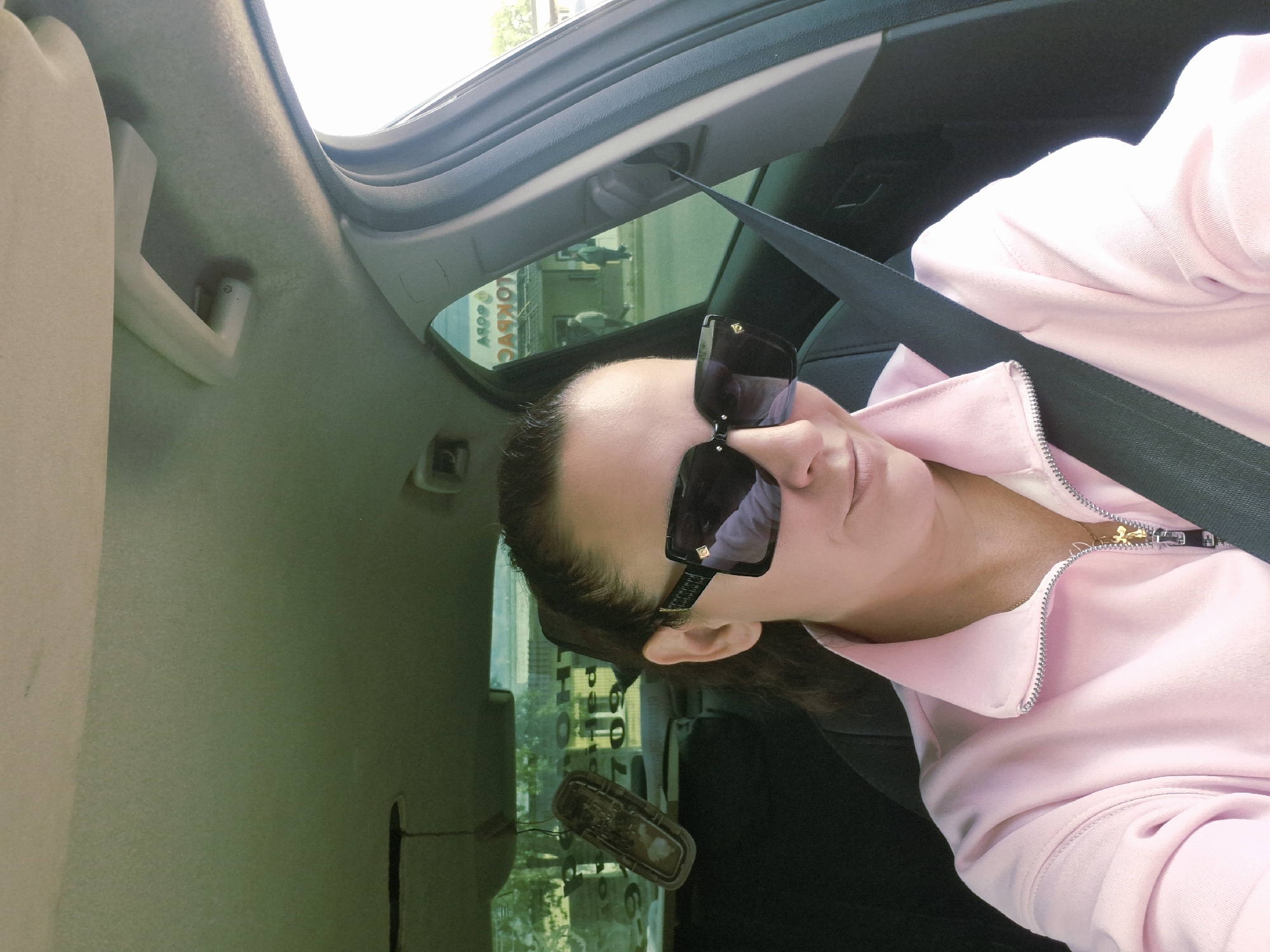 A woman is seated in a car, wearing a pink zip-up jacket and sunglasses. She is wearing a seatbelt. The car appears to be parked outside a building with signage visible through the window reflecting "МОТО ЗАПЧАСТИ", which translates to "Moto Parts". The location is in the city of Chaikovsky, Russia.