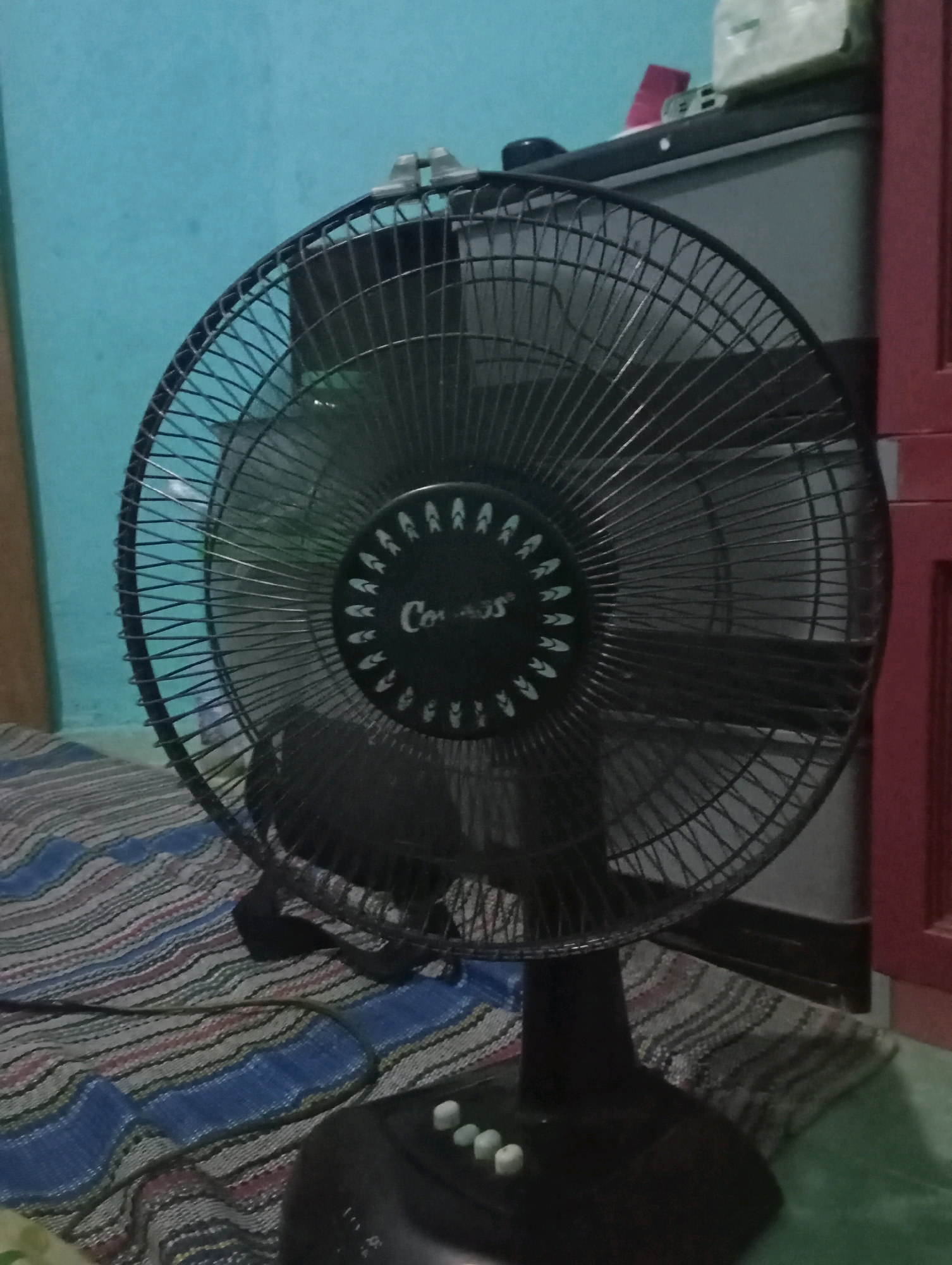 The image shows a black oscillating fan with a circular grill, positioned on a dark base. The fan is placed on a mat with a striped pattern of blue, white, and pink. In the background, a light blue wall is visible, along with the edge of a grey cabinet and a red cabinet. The scene appears to be an indoor domestic setting, possibly a bedroom or living area. No people are present in the image. The overall lighting is dim.