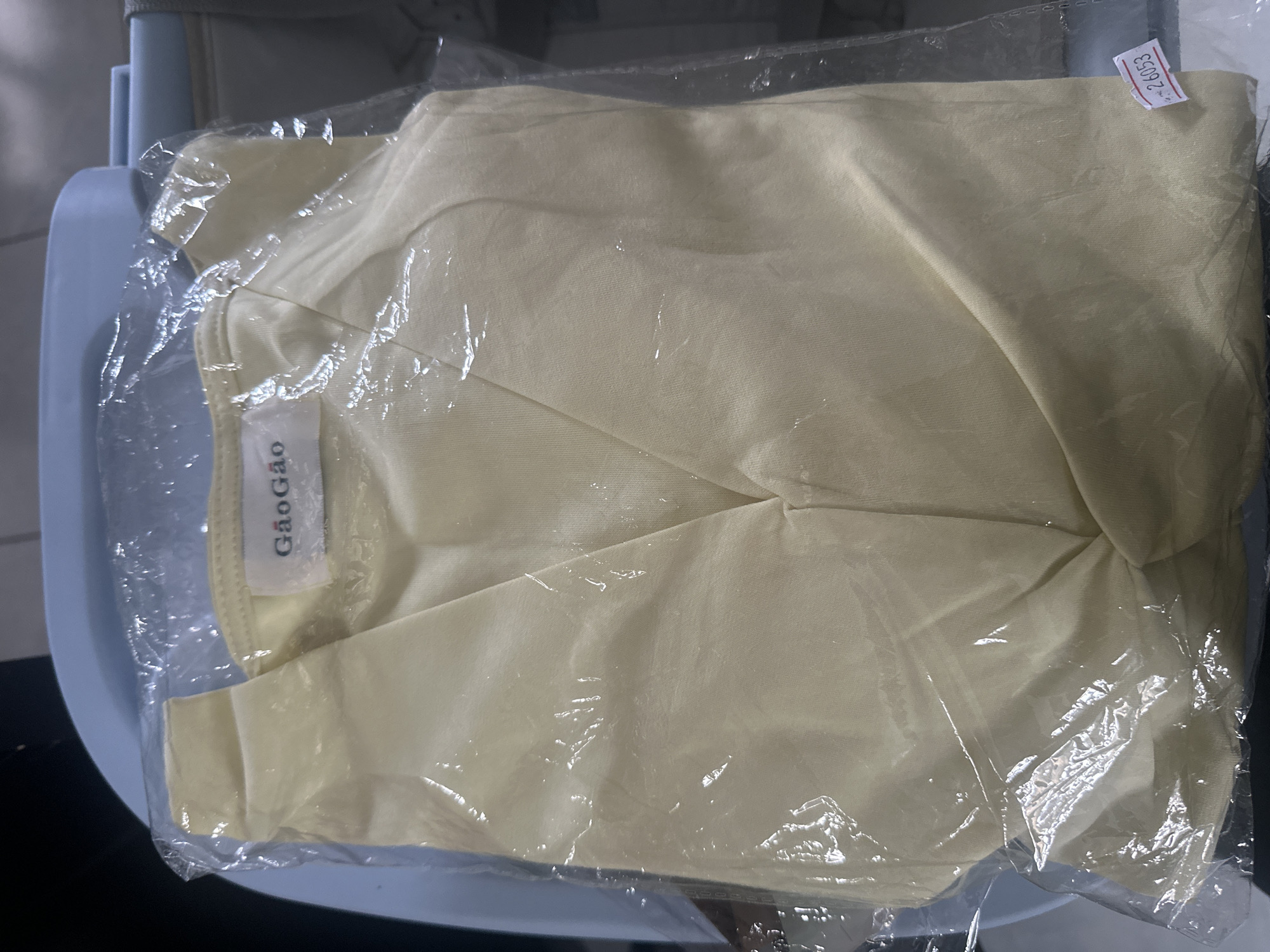 A pale yellow garment, possibly a blouse or dress, is presented inside a clear plastic bag, which is slightly wrinkled. A small white label with red lettering, reading "26053," is attached to the lower right corner of the plastic bag. The garment features a V-neckline and appears to be made of a smooth, possibly satin-like fabric. A tag with the brand name "GaoGao" is visible near the neckline. The item is placed on a light blue surface, likely a chair or a table, in an indoor environment. The lighting is overhead and casts subtle shadows.